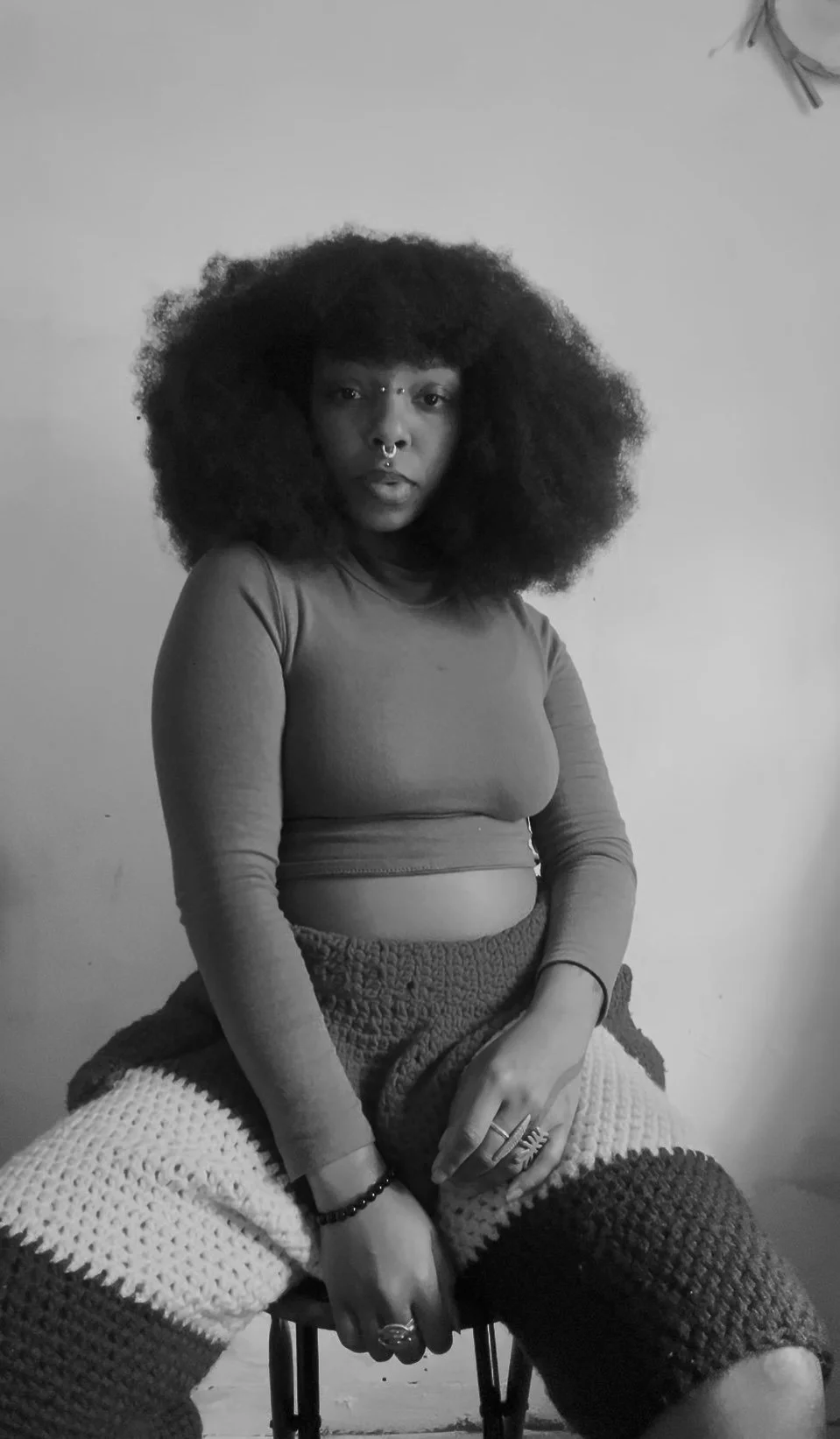 A woman with natural afro hair sitting on a chair against a plain wall, wearing a long-sleeve crop top and patterned pants, with rings and bracelets, looking directly at the camera.