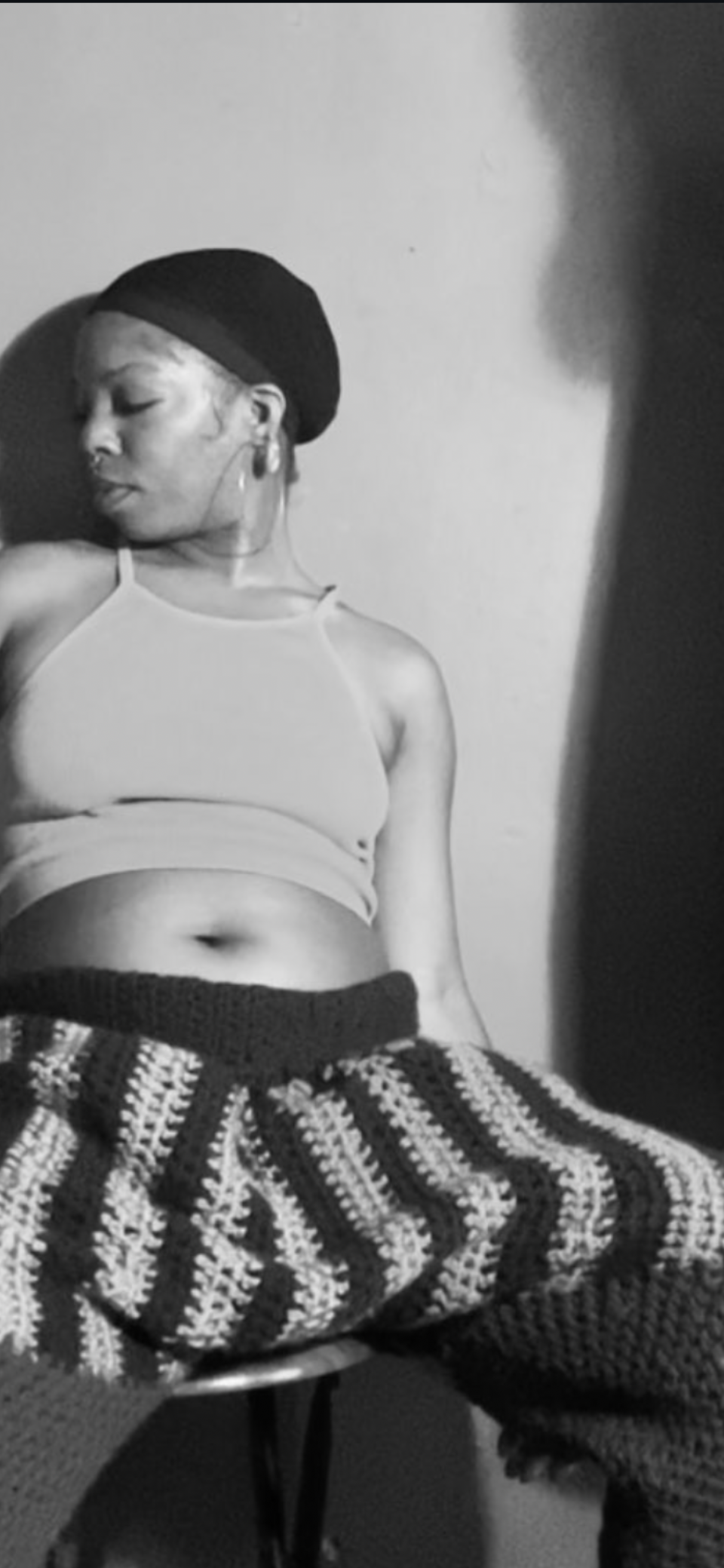 A woman sitting on a chair, wearing a sports bra and patterned pants, with her eyes closed and head tilted to the side, captured in black and white.