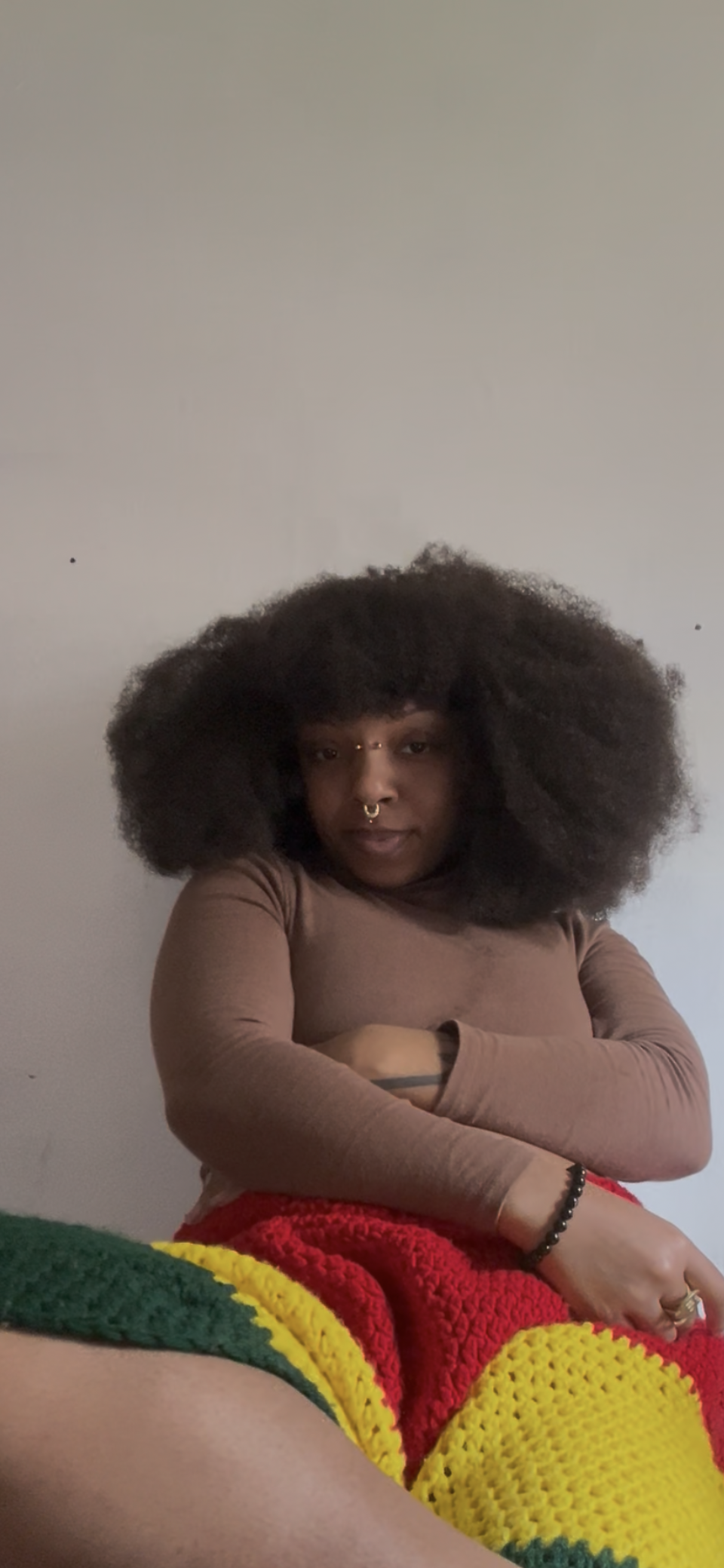A person with large curly hair, wearing a septum piercing, a brown long sleeve top, and a black bracelet, sitting on a colorful crochet blanket. The background is a plain white wall.
