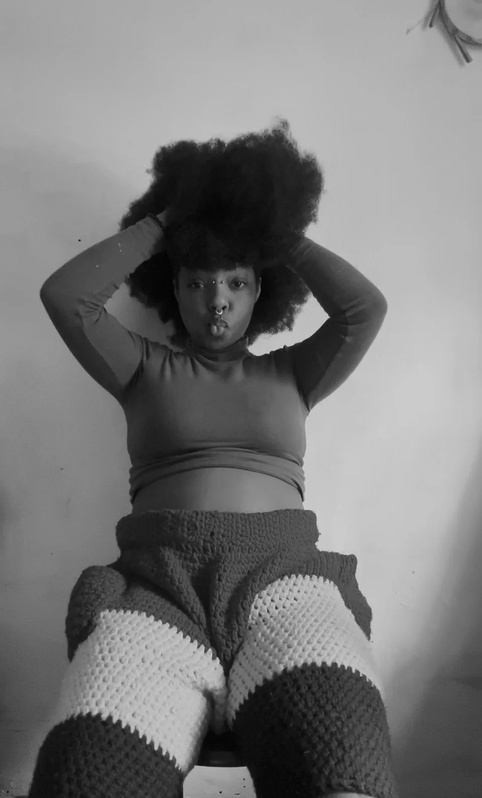 A woman with an afro hairstyle wearing a long-sleeve top and crocheted shorts, sitting and looking at the camera, in black and white.