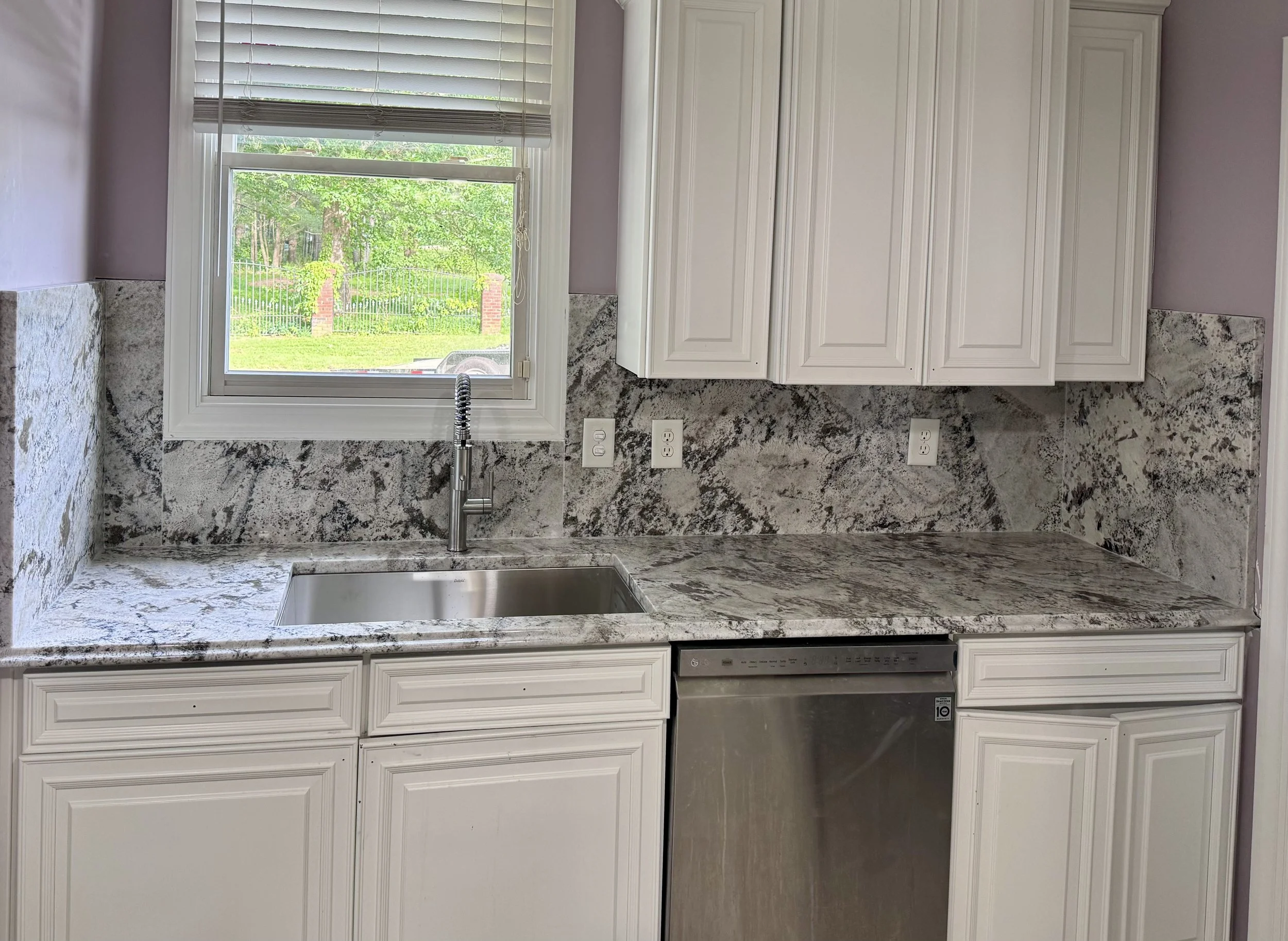 Kitchen with white cabinets, granite countertops, stainless steel dishwasher, single sink, window above the sink with view of green outdoor area.