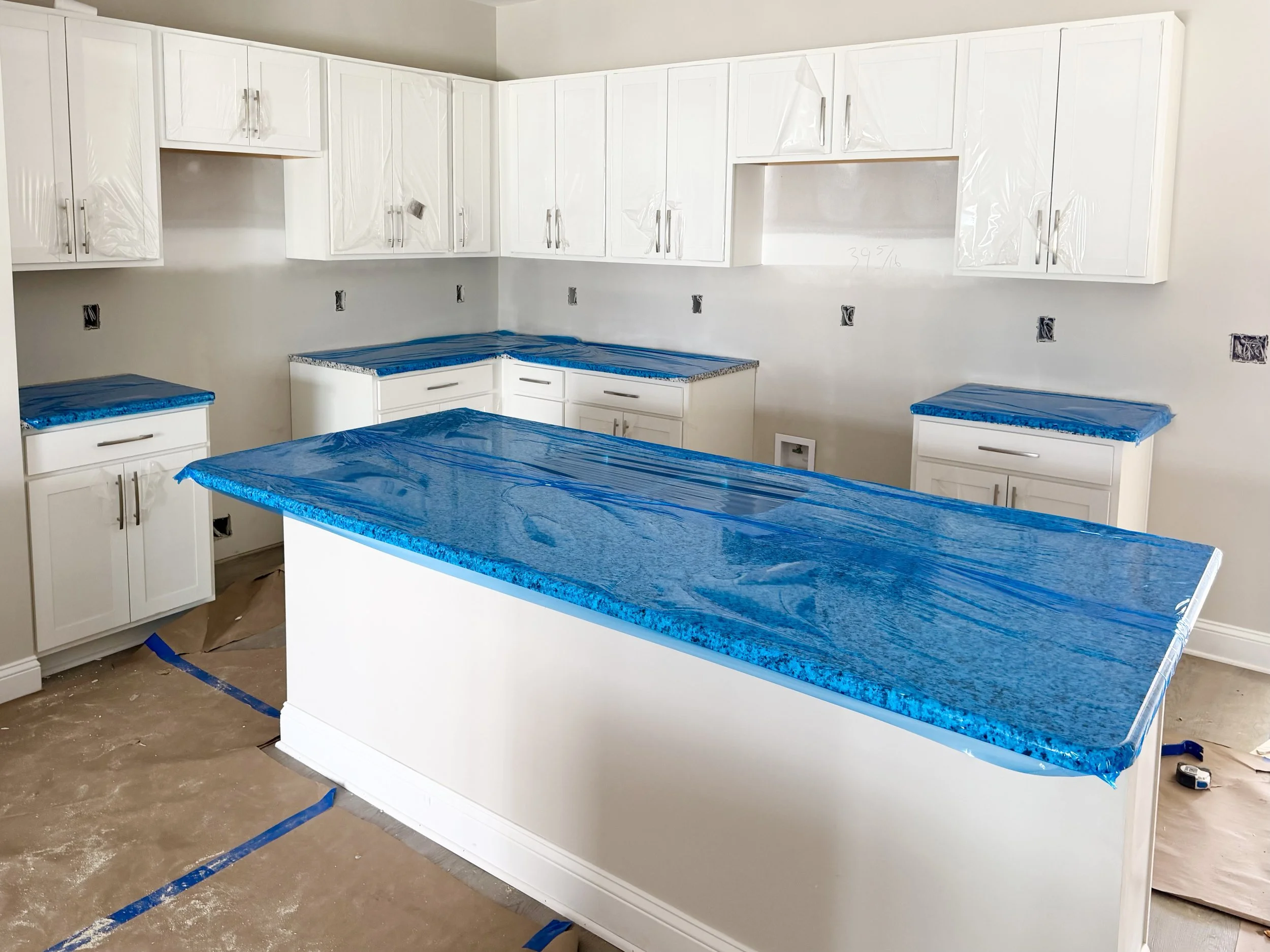 Kitchen under construction with white cabinets, some with protective plastic, a central island with a blue protective cover, and additional blue-covered surfaces on the counters and cabinets.