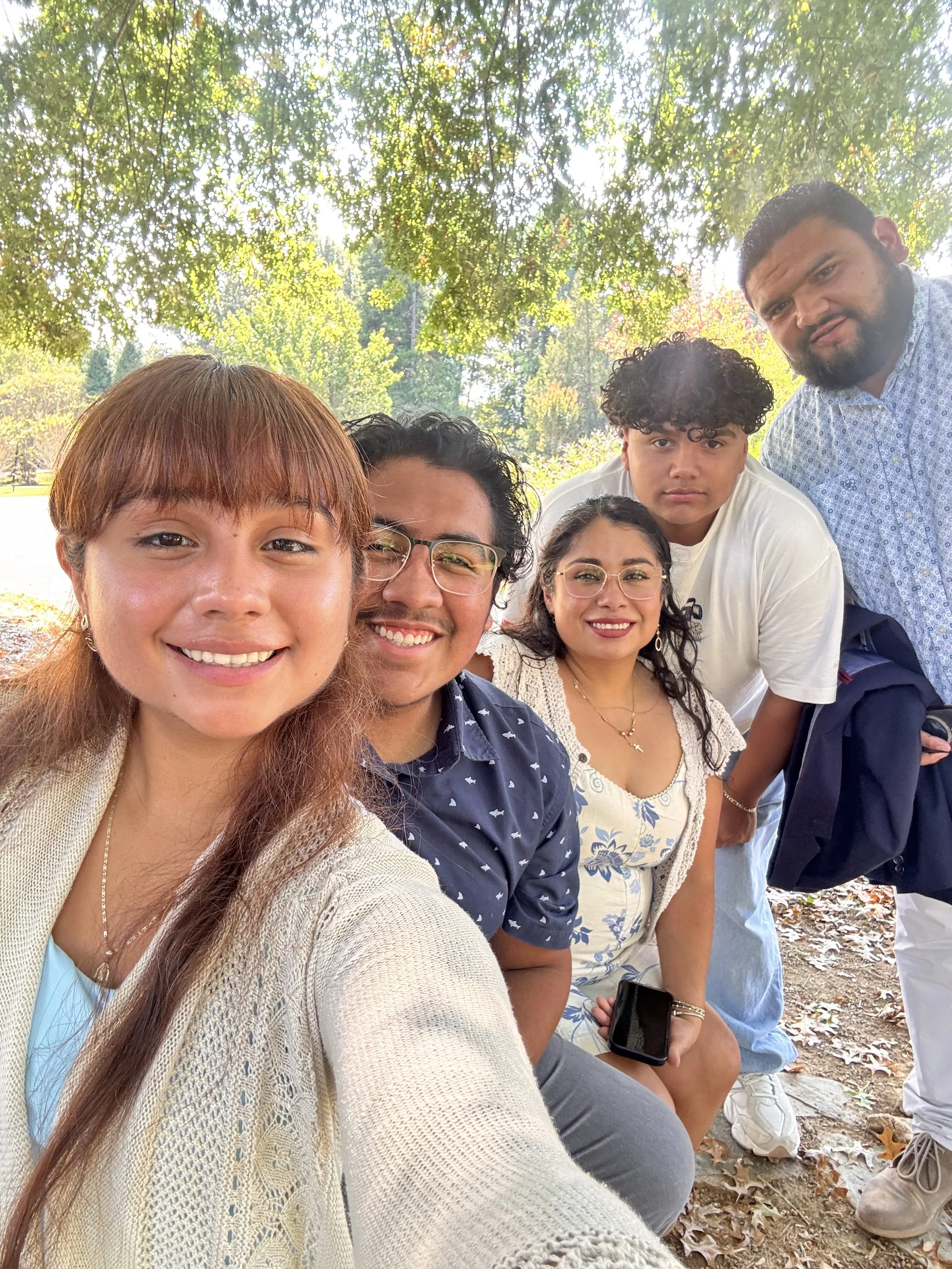 Group of six people taking a selfie outdoors under a large tree in a park with a pond and trees in the background, smiling and posing for the camera.