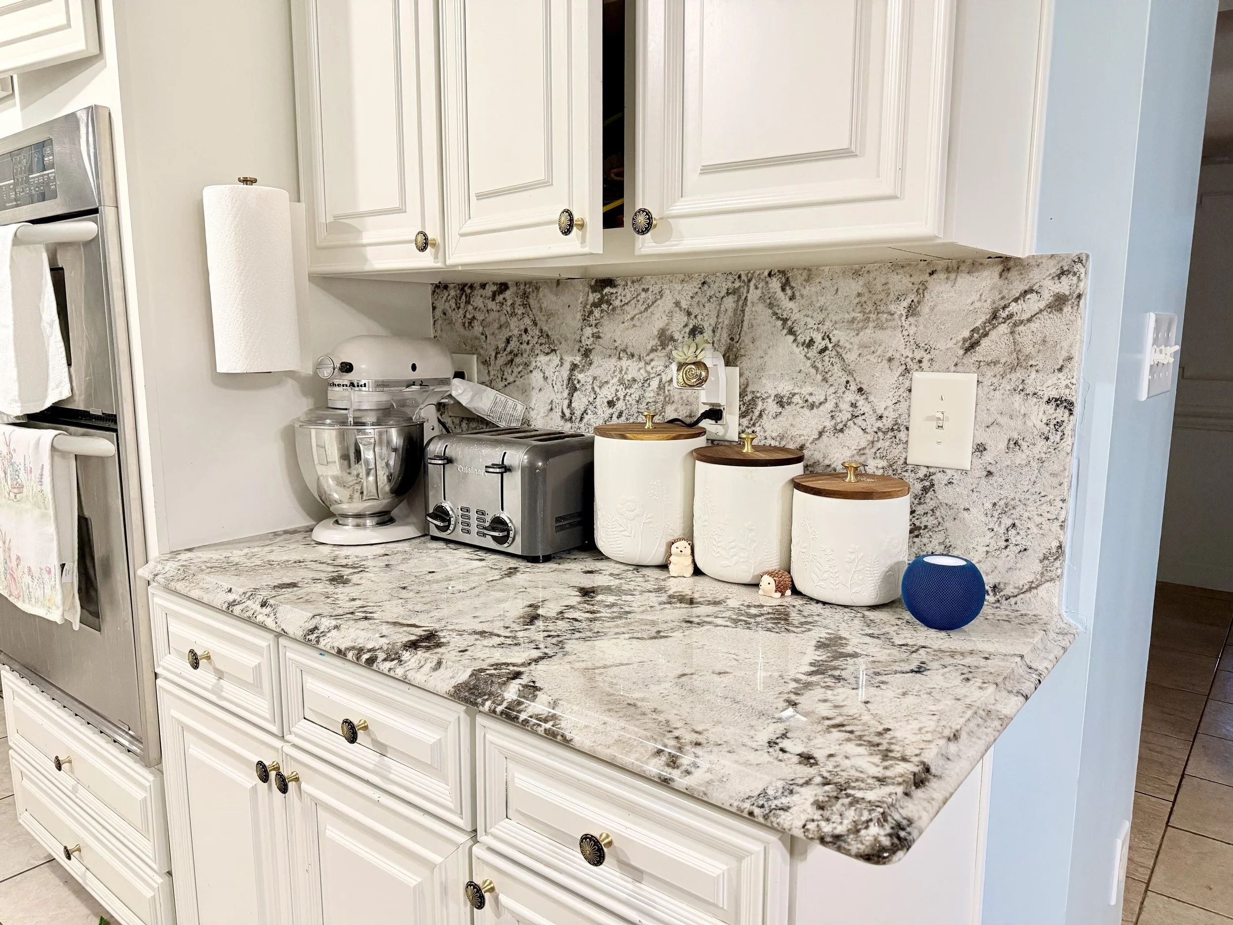 Kitchen countertop with a stand mixer, a toaster, three ceramic canisters with wooden lids, a small figurine, and a blue smart speaker. White cabinets above and white drawers below the granite countertop.