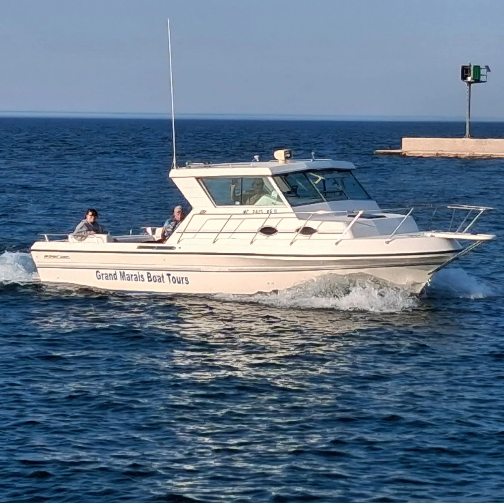 White boat named Grand Marais Boat Tours sailing on blue water with a small pier and navigational light tower in the background.
