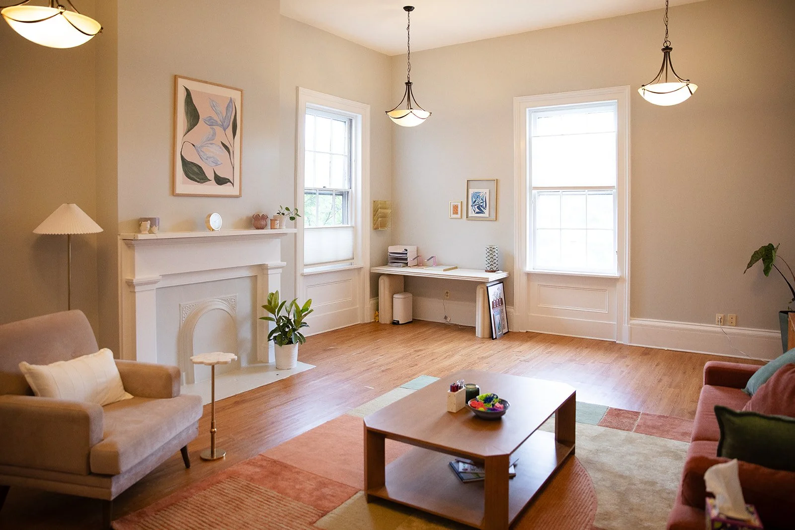 A bright living room with two large windows, light-colored walls, and hardwood floor. The room is furnished with a beige armchair, a pink sofa, a wooden coffee table with a bowl of colorful toys, and a desk against the wall with stacked papers. There are wall decorations, including a framed botanical print above the fireplace, and potted plants placed around the room.