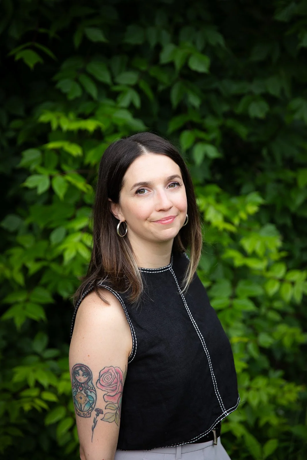 A woman with dark hair, wearing hoop earrings and a black sleeveless top, standing outdoors in front of green leafy foliage, with a tattoo of a girl, a rose, and a tree on her left arm.