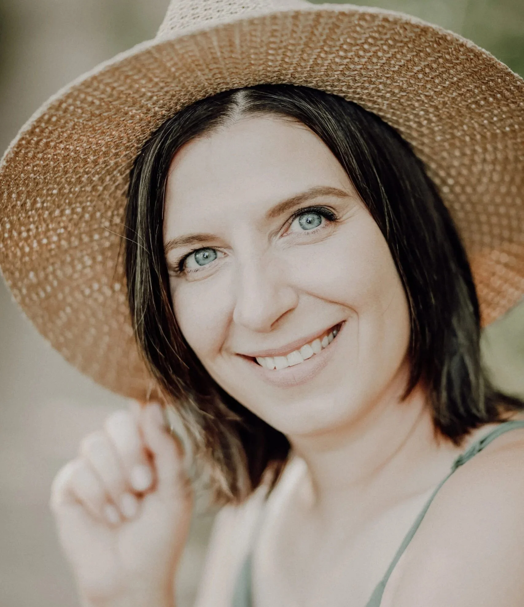Une femme souriante portant un chapeau en paille, avec des yeux bleus et des cheveux noirs, dans un cadre naturel.