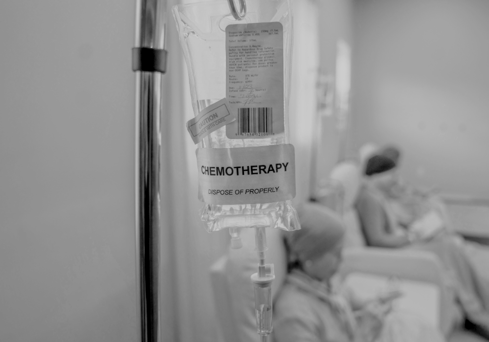 Une perfusion de chimiothérapie suspendue dans un hôpital, avec des patients assis dans des lits en arrière-plan.