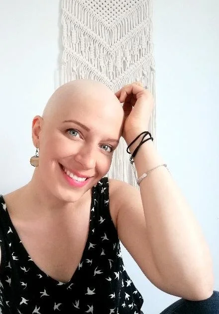 Une femme souriante, sans cheveux, portant un haut noir avec des motifs d'oiseaux, de bijoux, devant un décor avec un motif en macramé blanc