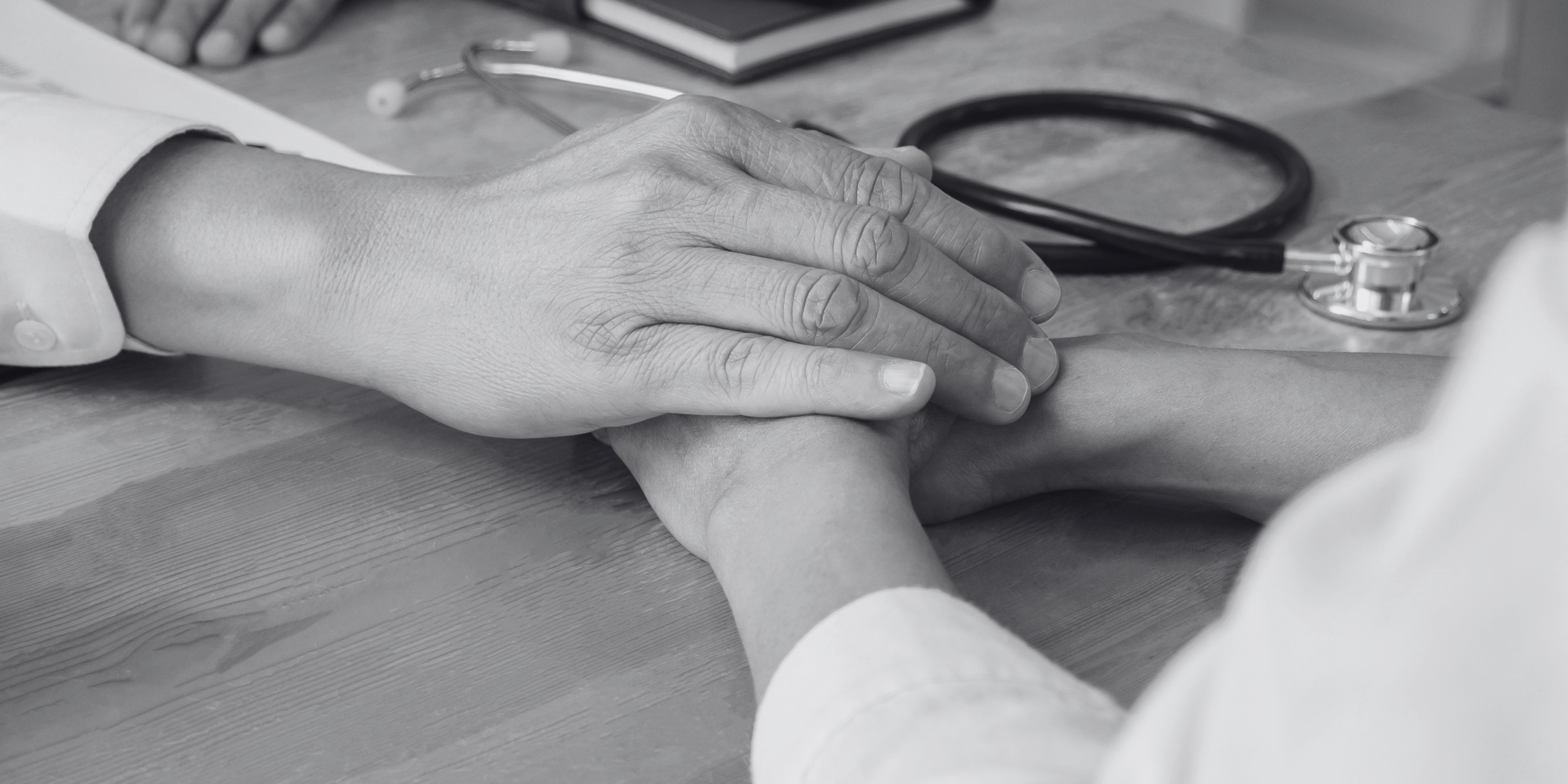 Une main d'un médecin tenant la main d'un patient sur une table en bois, avec un stéthoscope en arrière-plan.