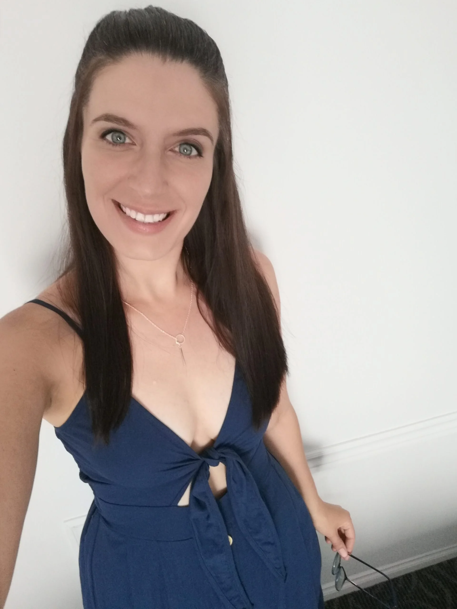 Une femme souriante avec des cheveux bruns longs, portant une robe bleue avec un nœud à la taille, prise en selfie dans une pièce avec un mur blanc.