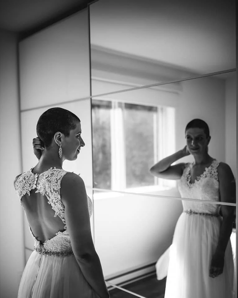 Une femme aux cheveux rasés portant une robe de mariée avec un décolleté en dentelle regarde son reflet dans un miroir, retrouvant son image dans un miroir mural, dans une pièce lumineuse avec une fenêtre derrière.
