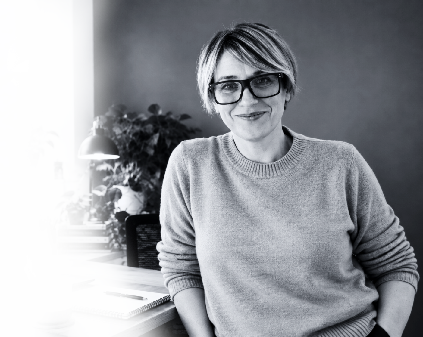 Black and white photo of a woman with short hair, glasses, and a sweater, sitting in an office with plants and a desk with a notebook, pen, coffee mug, and laptop.