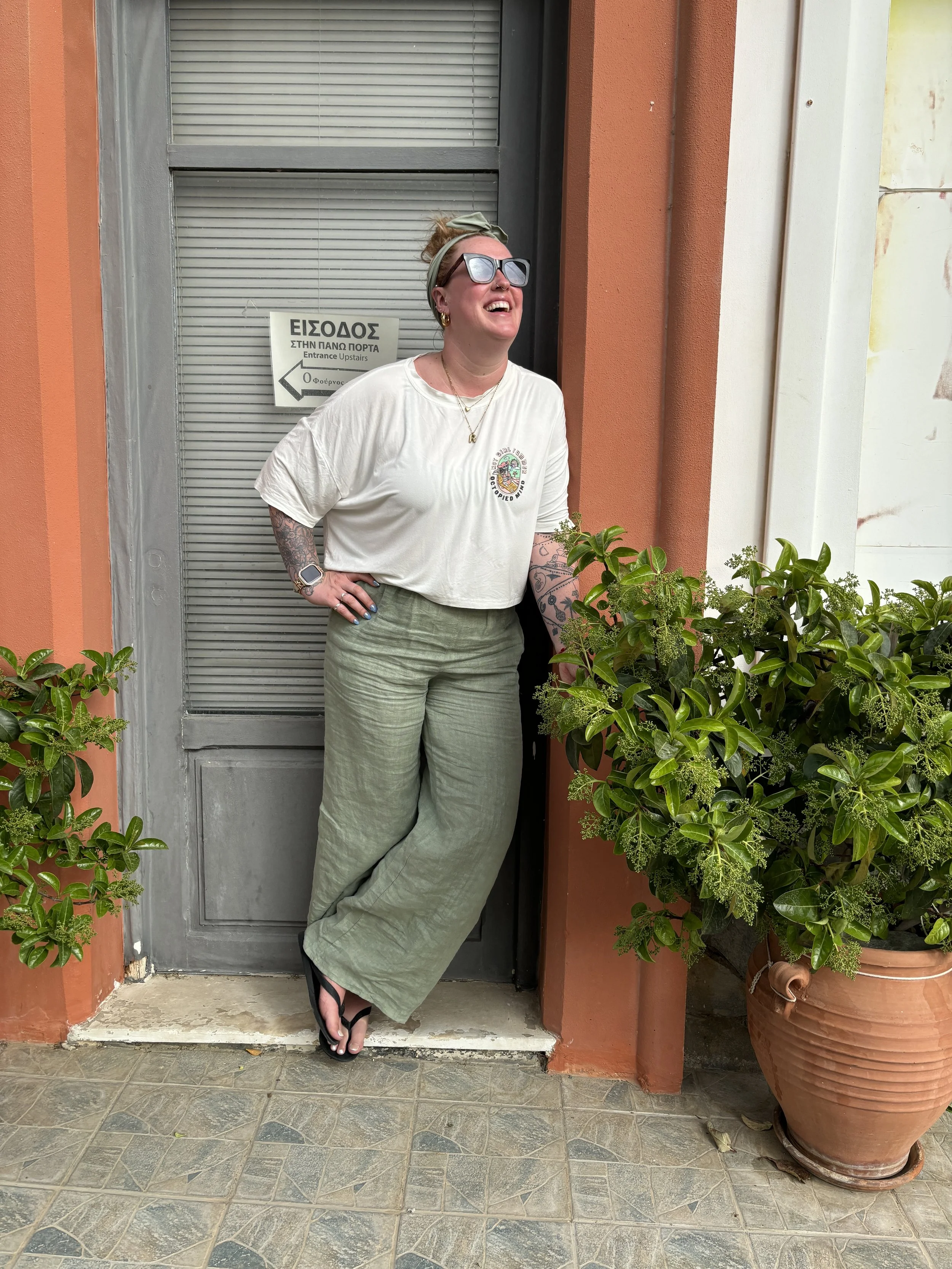 A woman standing outdoors next to a large potted plant in front of a gray door with a sign. She is wearing sunglasses, a white T-shirt, green wide-leg pants, and flip-flops. She is smiling and looks happy.