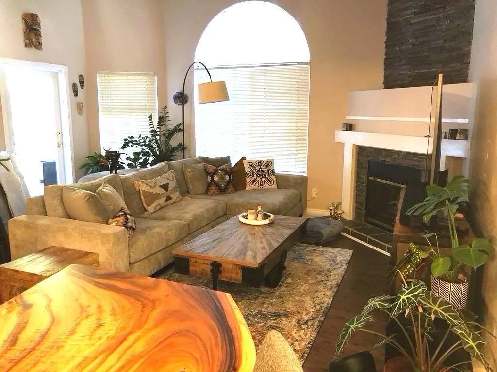 Living room with beige sofa, wooden coffee table, plants, fireplace, window blinds, colorful pillows, and decorative items.
