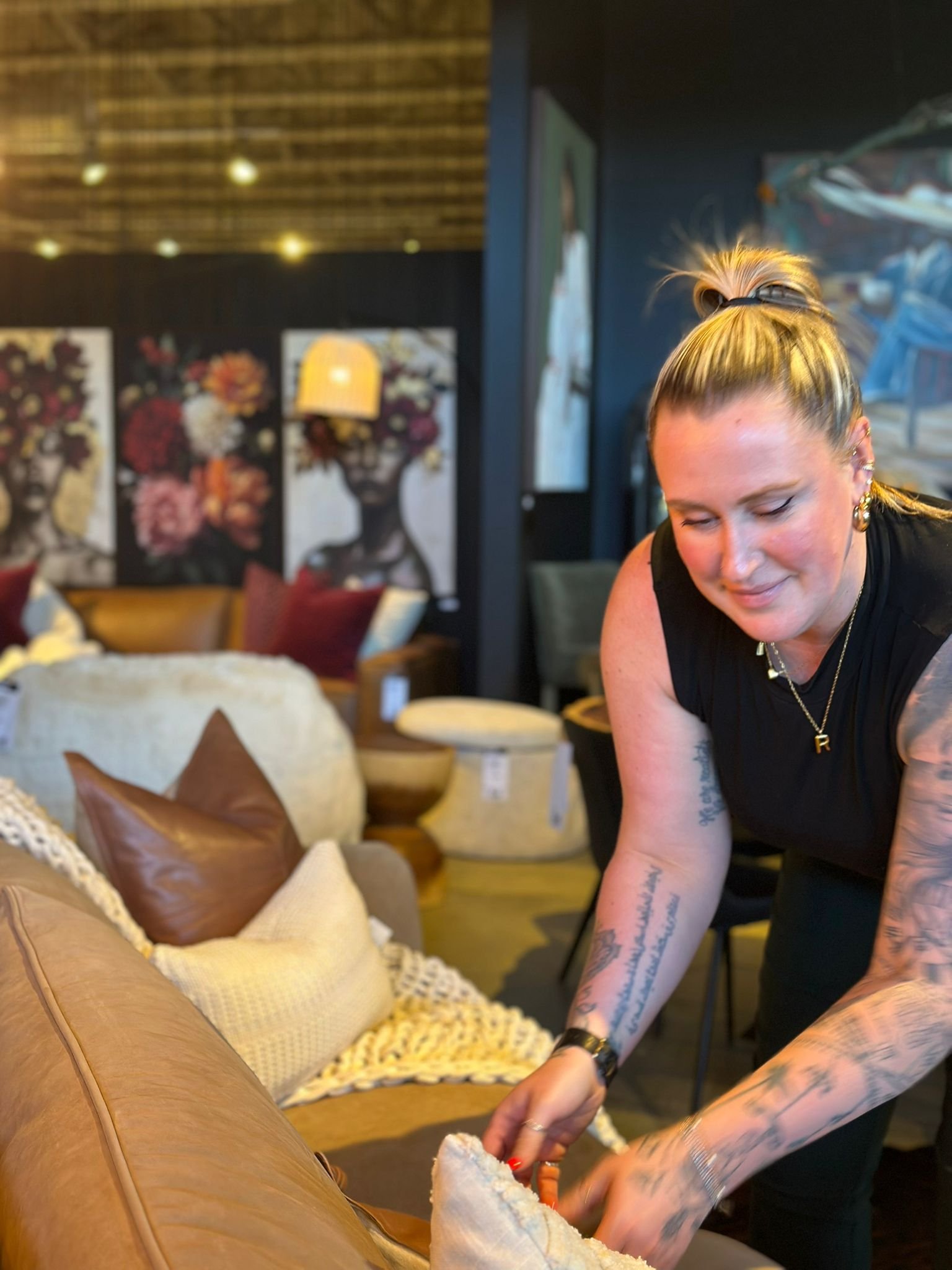 A woman with blonde hair tied in a ponytail and tattoos on her arms is leaning over a brown leather couch in a decorated living room, arranging or adjusting pillows and a blanket on the couch.