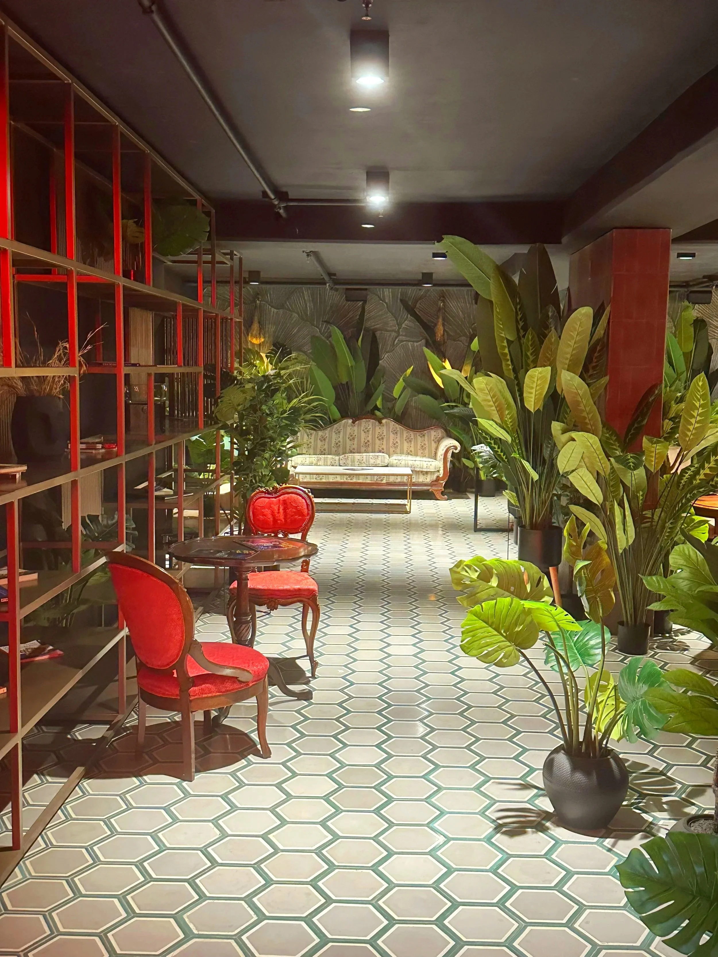 Interior of a room with patterned tile floor, large potted plants, a vintage sofa, and red velvet chairs, with overhead lighting and a bookshelf on the left.
