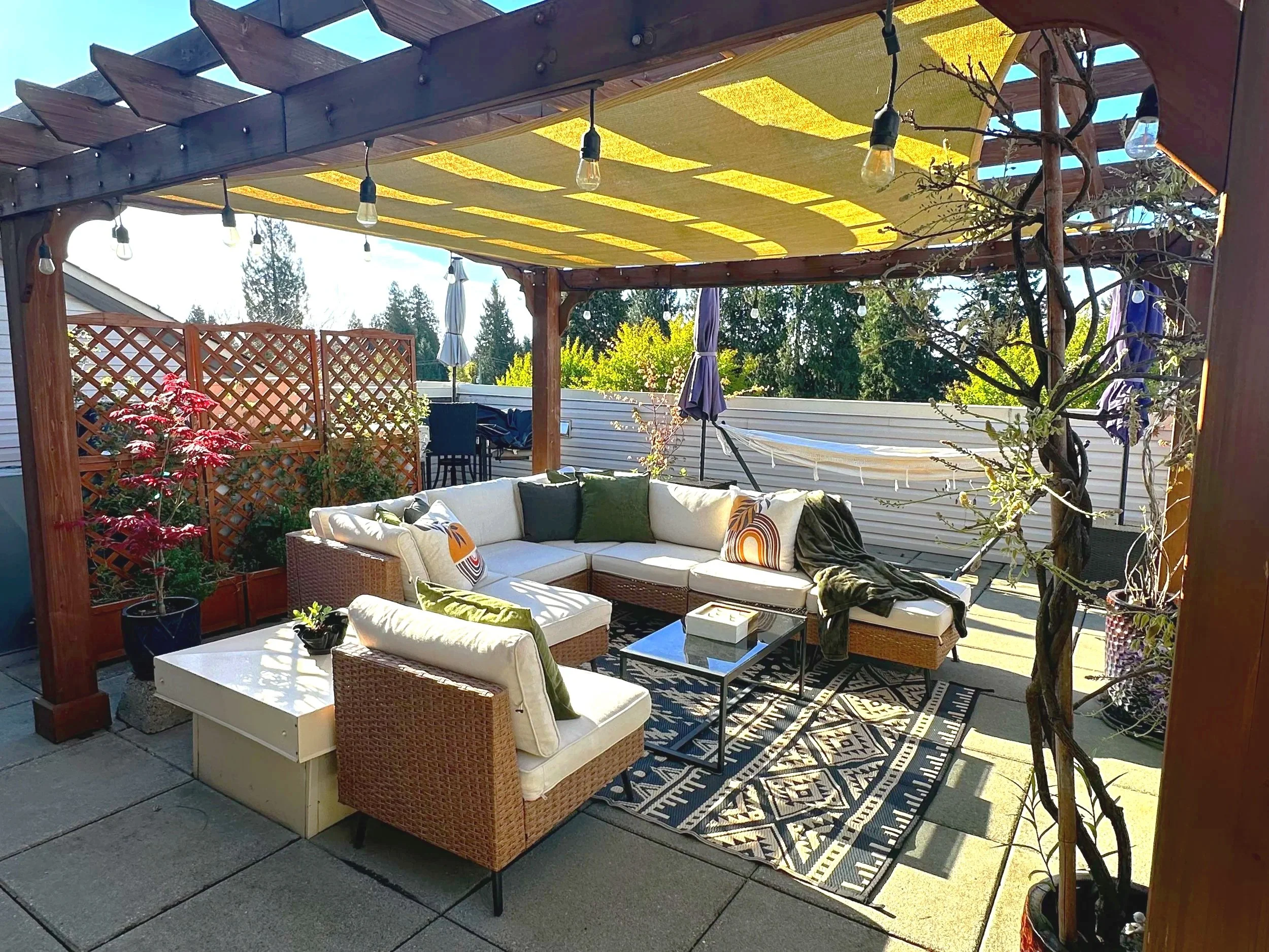 Outdoor patio with a pergola covered with yellow fabric, string lights hanging from the beams, seating area with a white sectional sofa with green and patterned cushions, a wicker chair, a small black metal table, plants in pots, a rug with geometric pattern, and umbrellas in the background, with trees and sky visible beyond the fencing.
