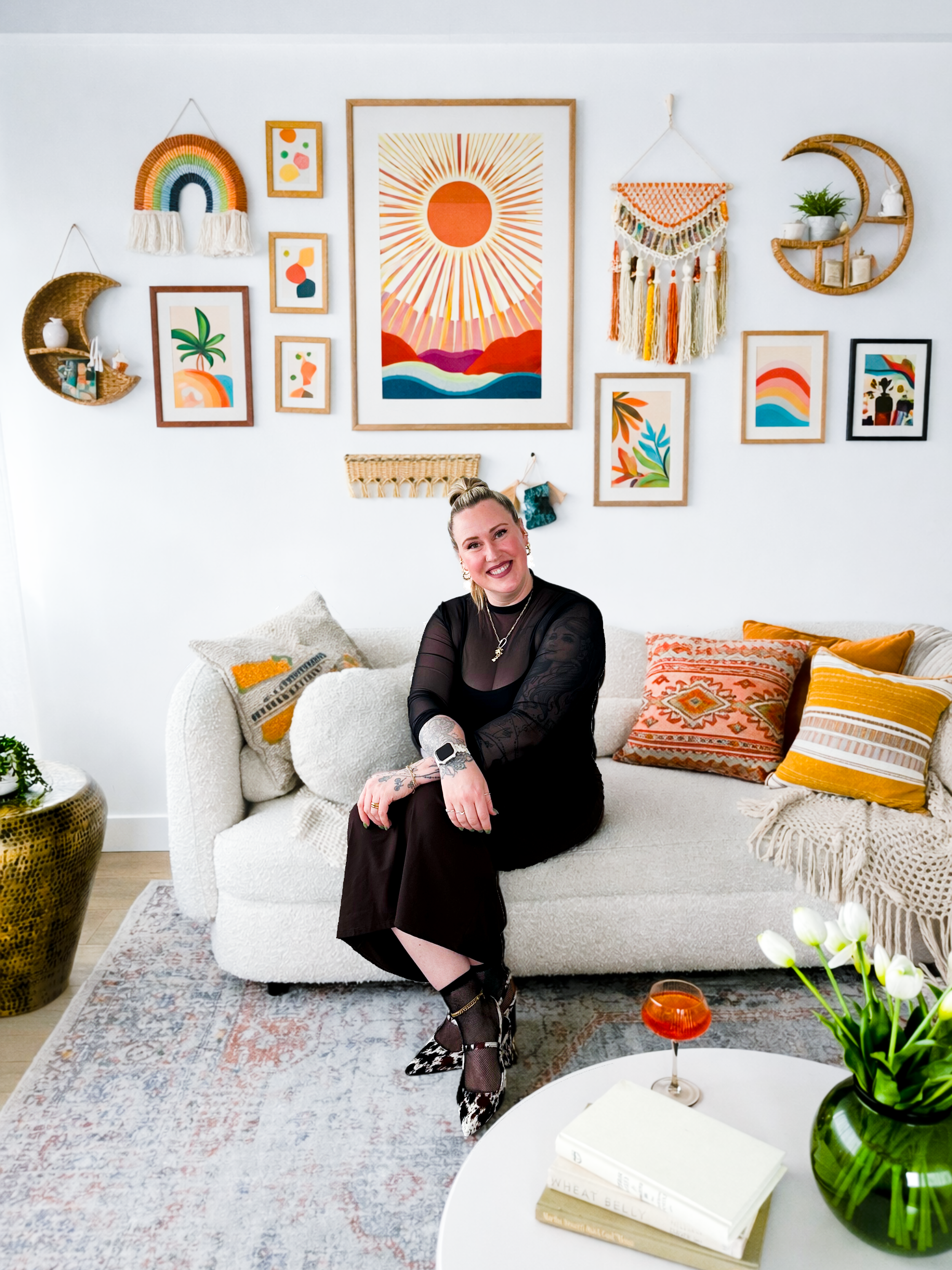 A woman sitting on a white sofa in a living room with colorful wall art and decor. She is smiling, wearing a black outfit, and has tattoos on her arm. There are patterned pillows on the sofa, a round coffee table with books and flowers, and a large green vase on a side table.