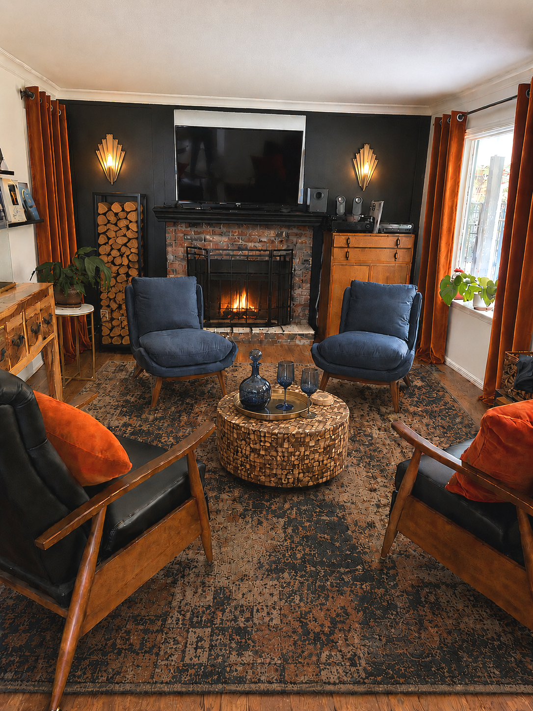 Living room with a brick fireplace, two blue armchairs, a black sofa with orange pillows, a round patterned ottoman with blue glassware, a patterned rug, and a large television above the fireplace. Window with orange curtains on the right and greenery on the window sill.