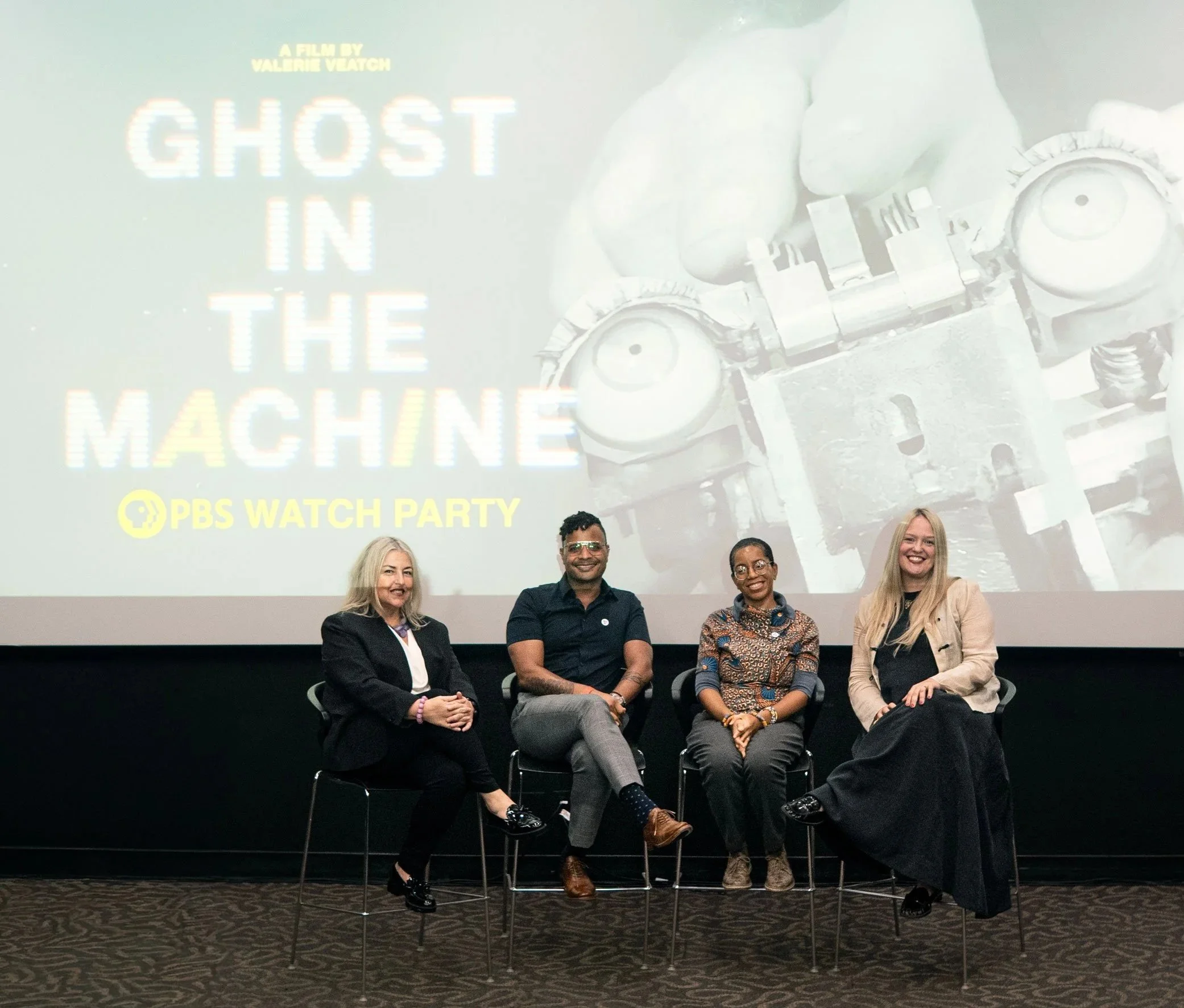Erica Chito Childs and three other people sitting on chairs on stage in front of a large screen. The screen displays the PBS logo and the title 'Ghost in the Machine' with a subtitle 'A Film by Valerie Veatch' and the PBS Watch Party logo.
