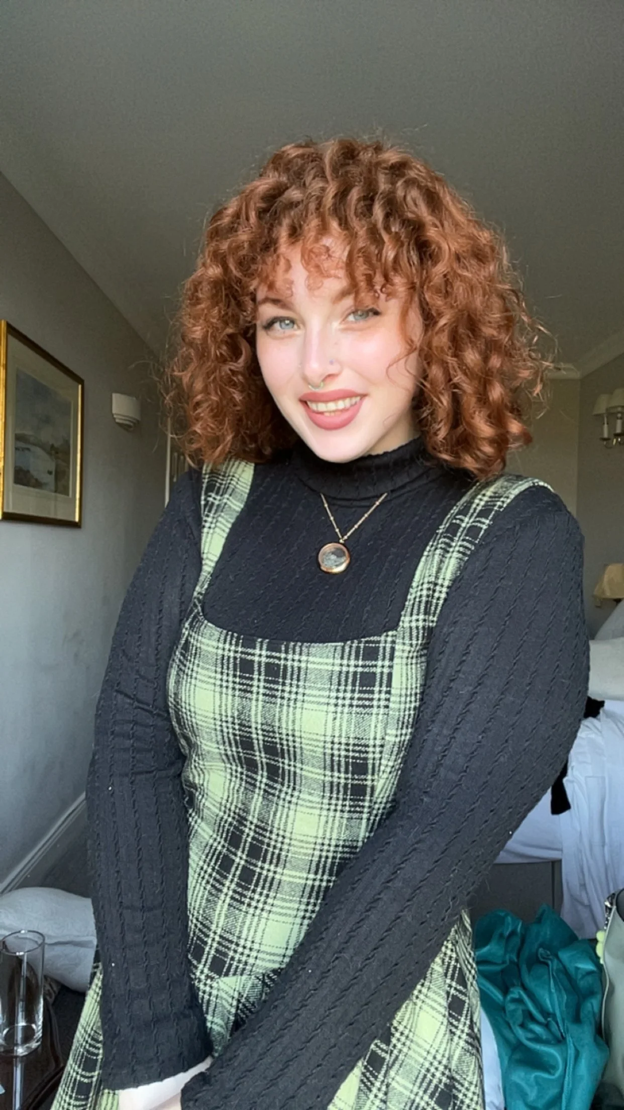 A woman with curly red hair smiling, wearing a black turtleneck, a plaid green and black dress, and a pendant necklace, standing indoors in a hotel room or bedroom.