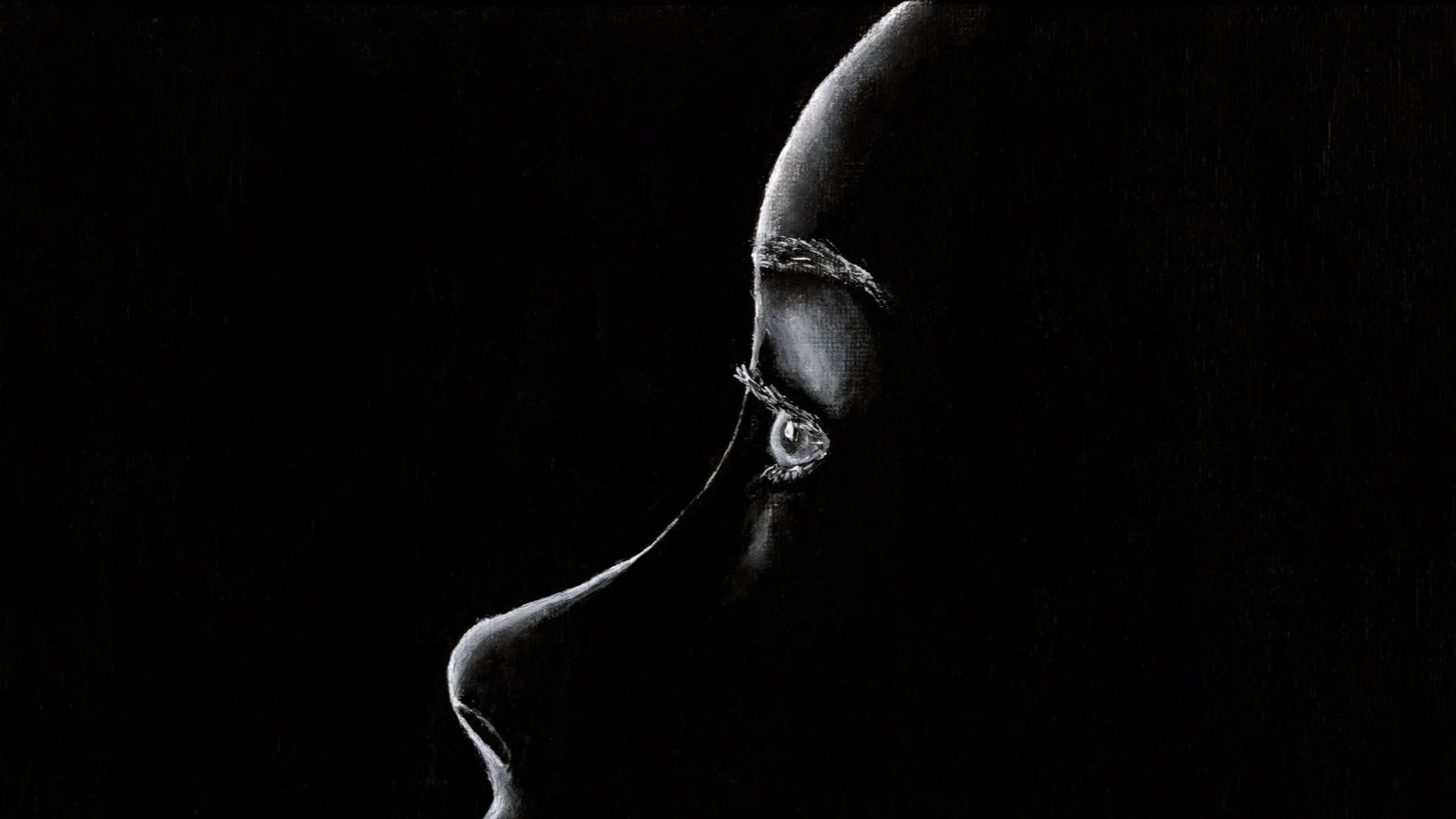 A dark, monochrome profile of a human face in shadow, showing only the silhouette and features such as the nose, lips, and eye socket.