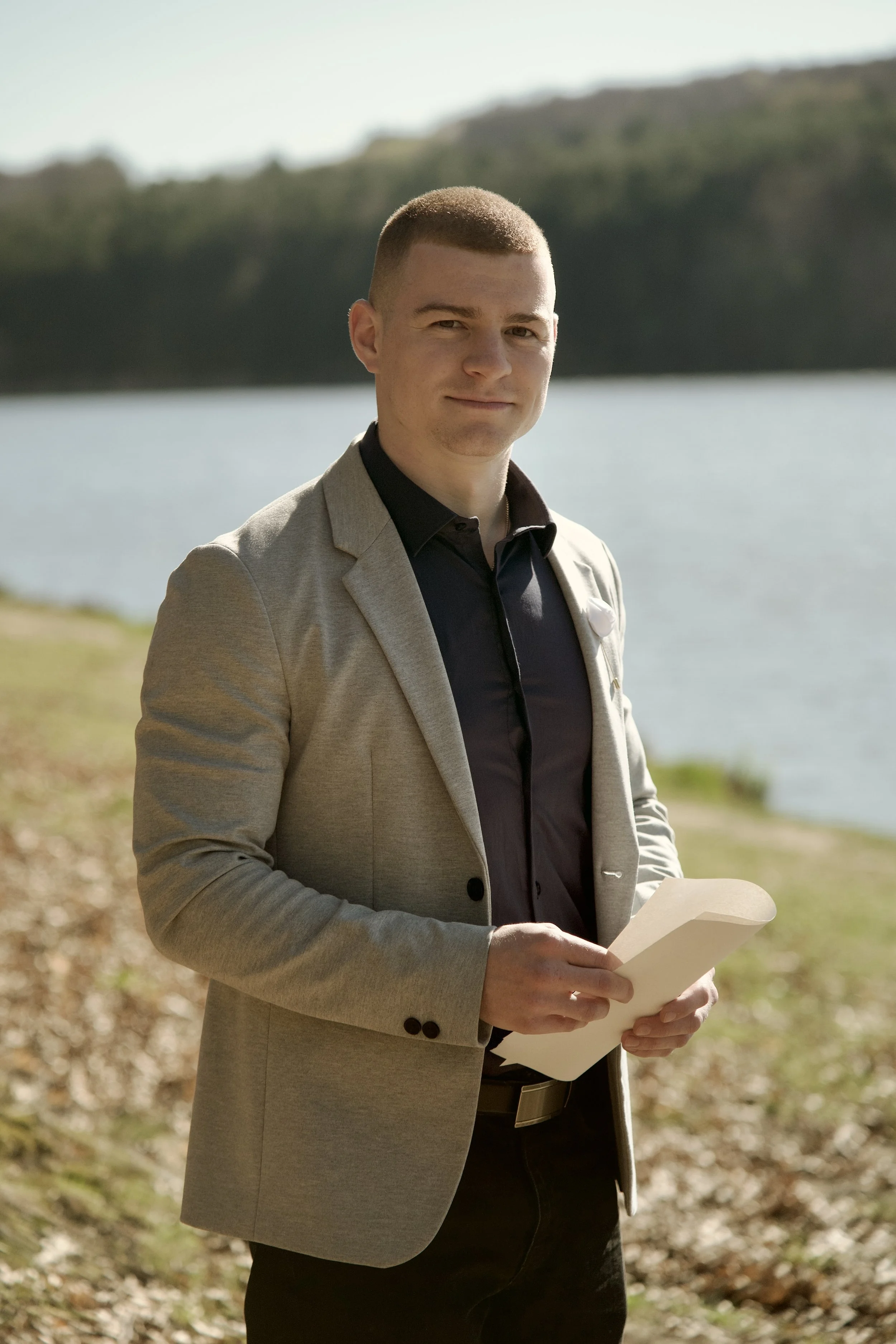 Egy szürke zakót és sötétkék inget viselő férfi ceremóniamester áll a szabadban egy vízparti esküvőn, egy jegyzettömböt tartva a kezében.