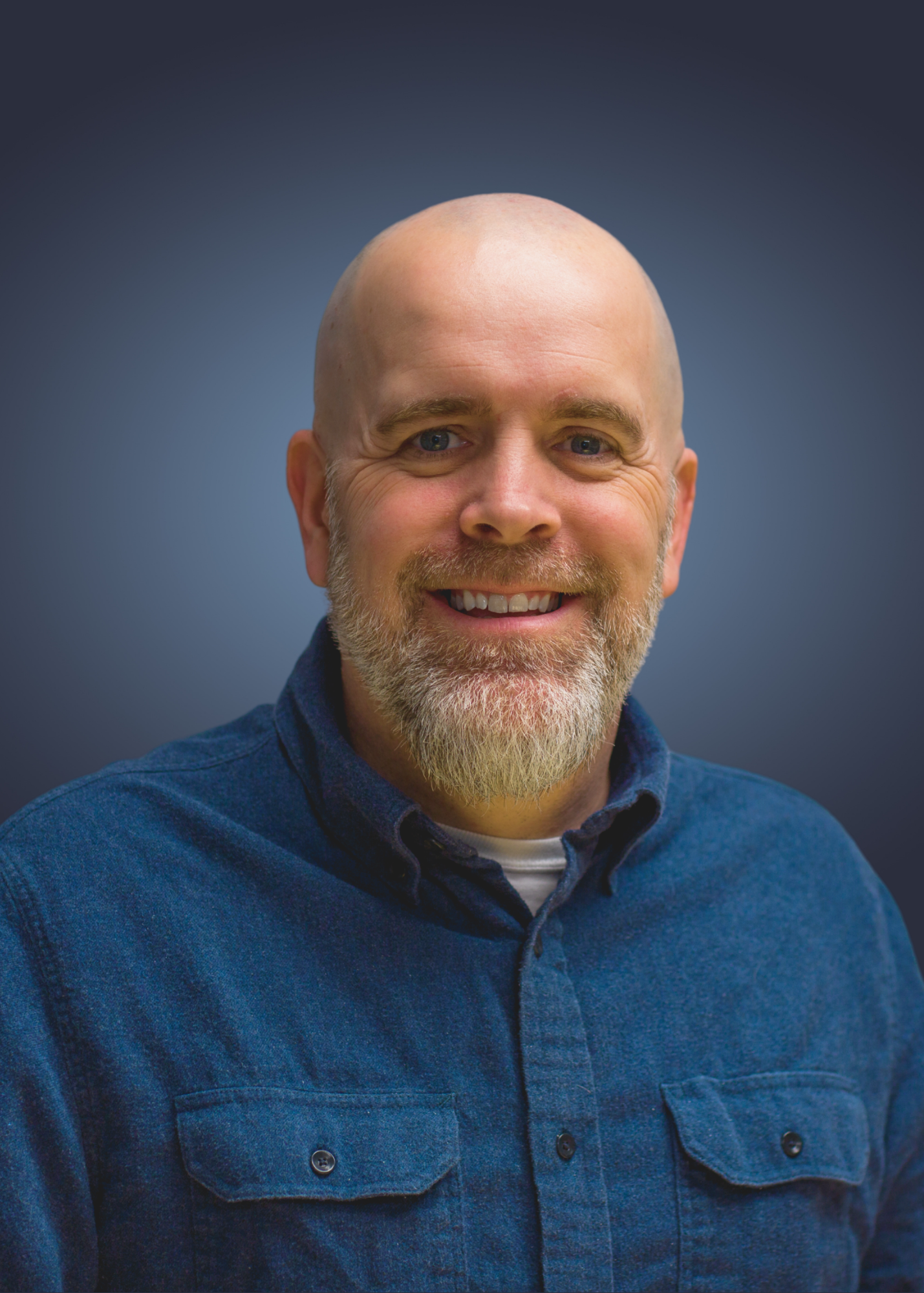 Headshot of the founder Ryan Worley with a beard, wearing a blue button-up shirt, standing against a blurred blue background.