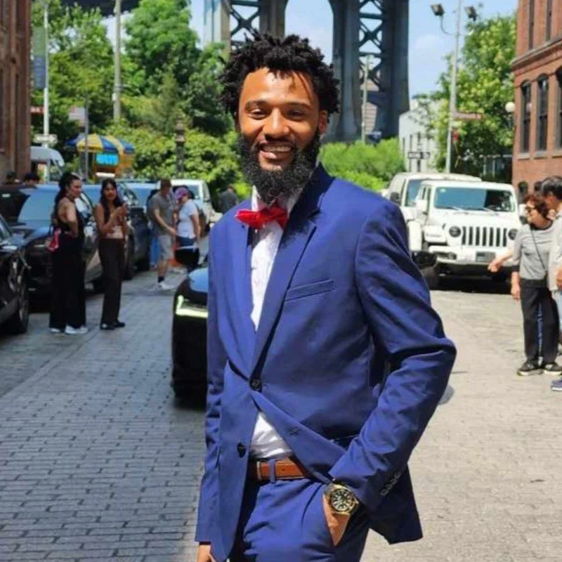 Man with curly hair and a beard, wearing a blue suit, red bow tie, and white shirt, smiling outdoors on a city street with people, cars, and a bridge in the background.