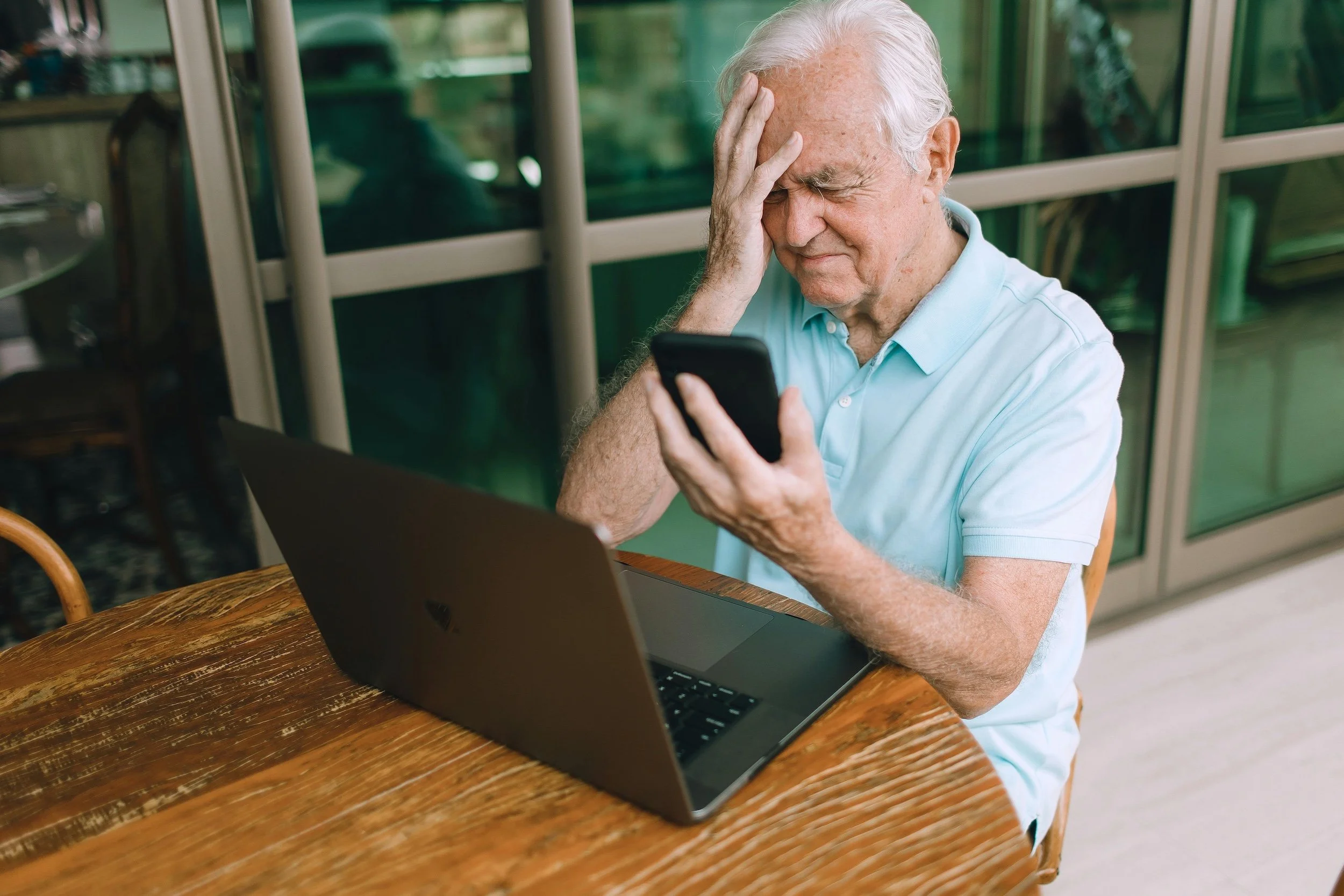 An elderly man with white hair wearing a light blue polo shirt is sitting at a wooden table with a laptop in front of him, looking at his smartphone with a distressed expression, holding his head with one hand.