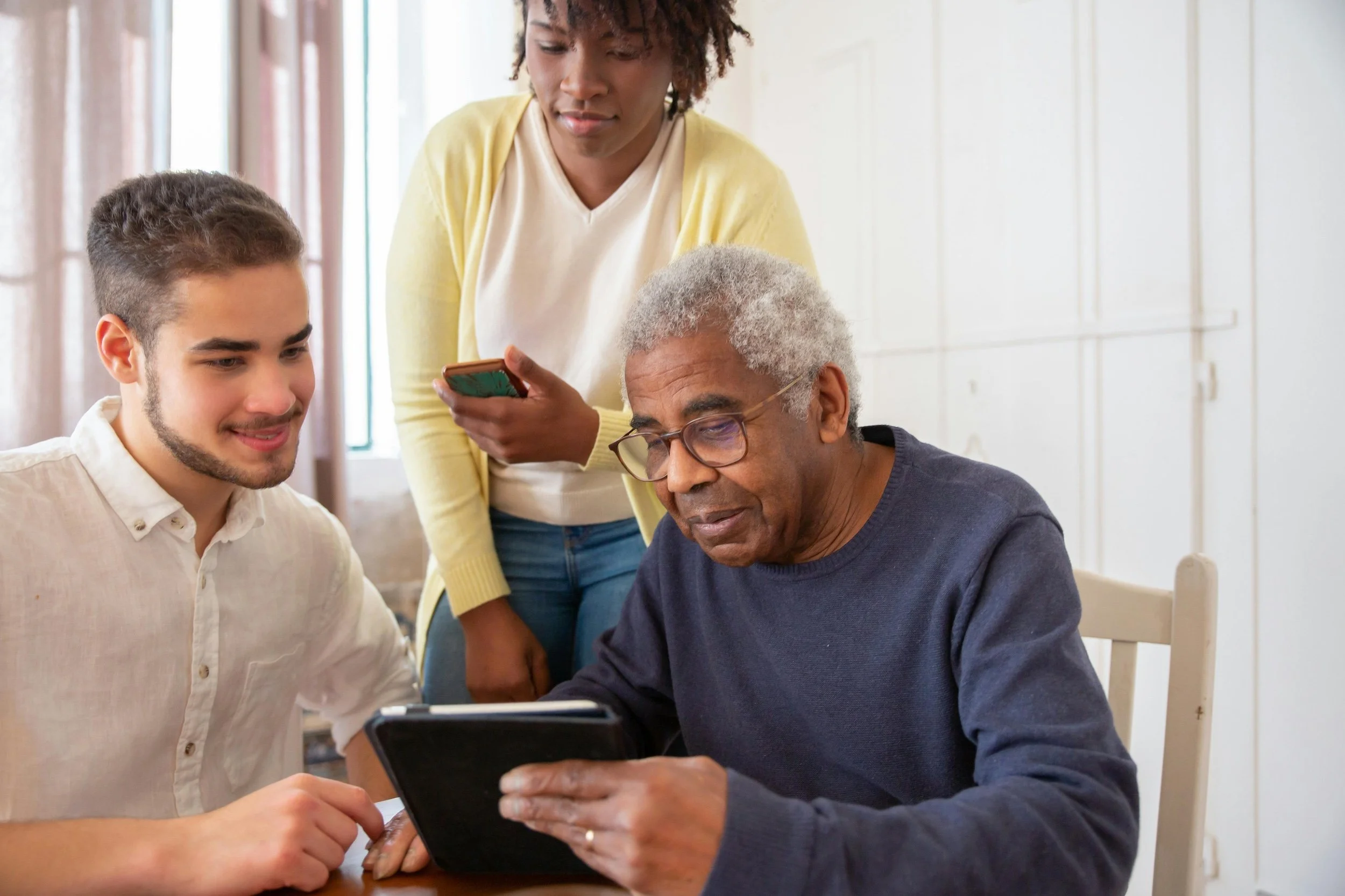 Three people gathered around an older man using a tablet, while a woman stands behind them looking at her phone.