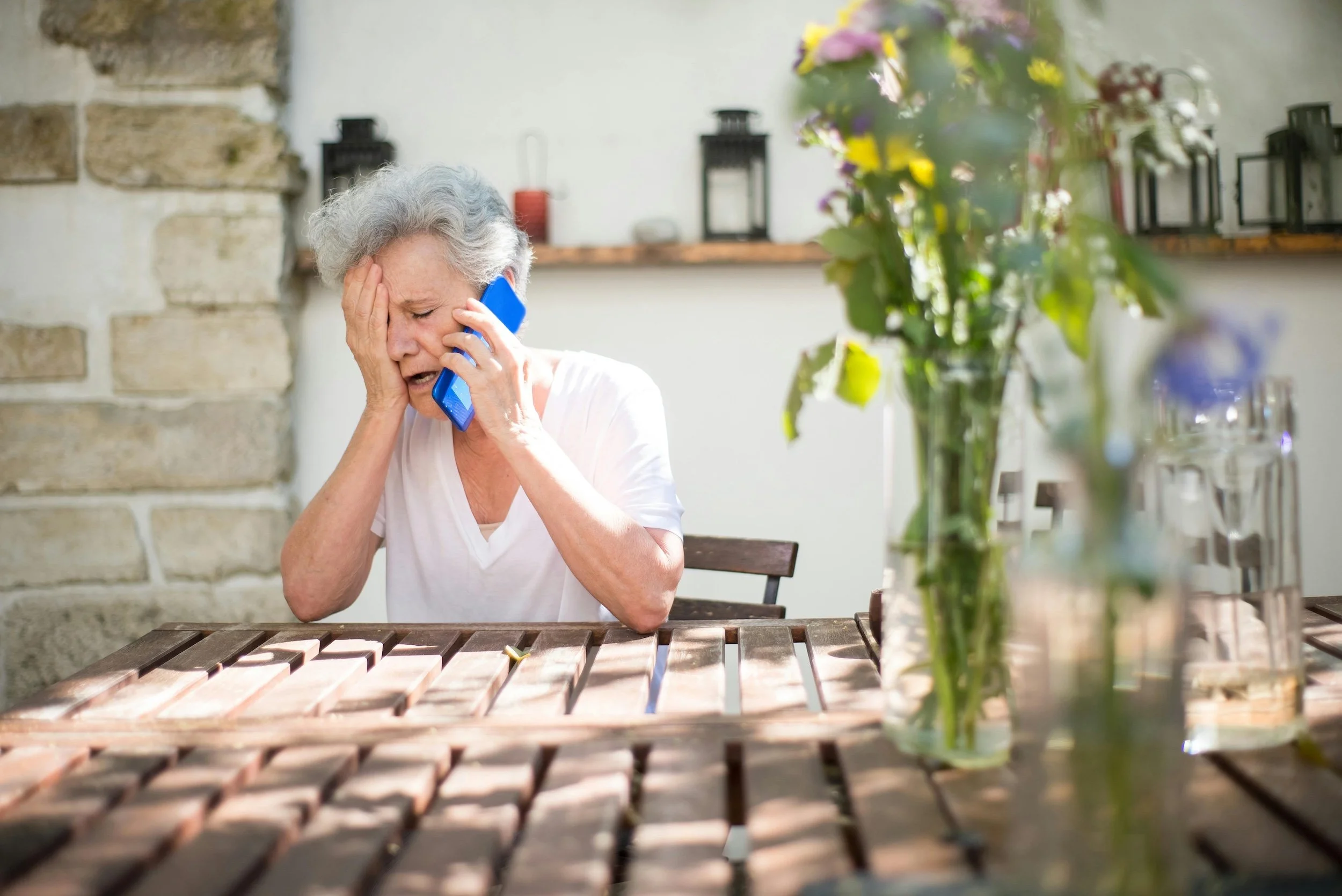 An elderly woman with gray hair sitting at a wooden table outside, holding her head with one hand and a blue phone to her ear with the other, appearing distressed.