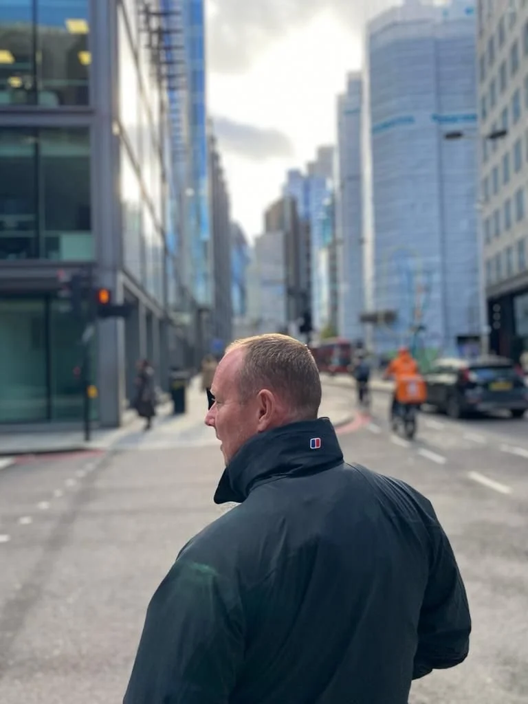 A man in a black jacket walking down a city street with tall buildings on either side