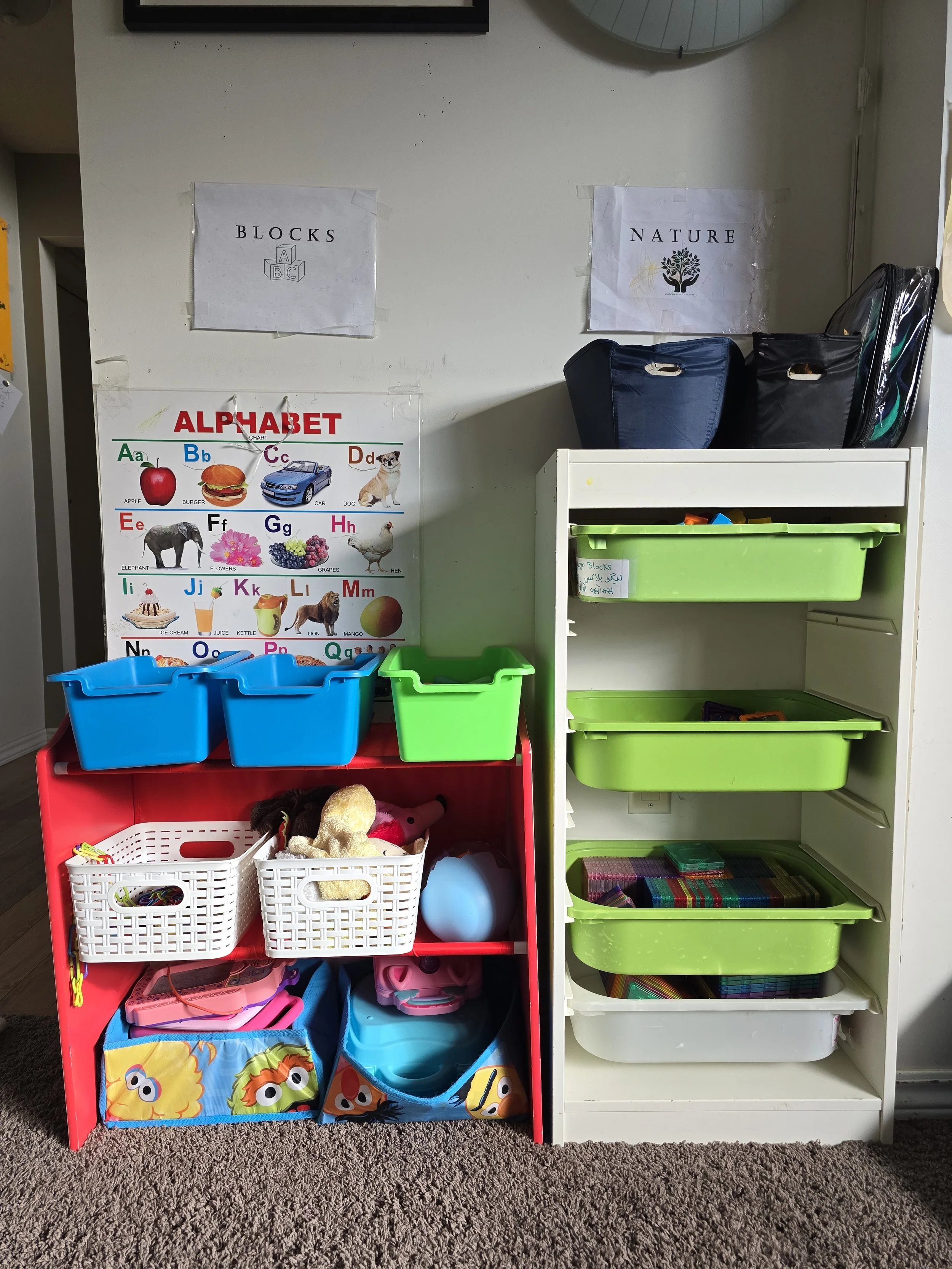 Organized children's educational and play supplies including bins with toys, books, and learning materials, with alphabet chart and labeled storage on white shelves.