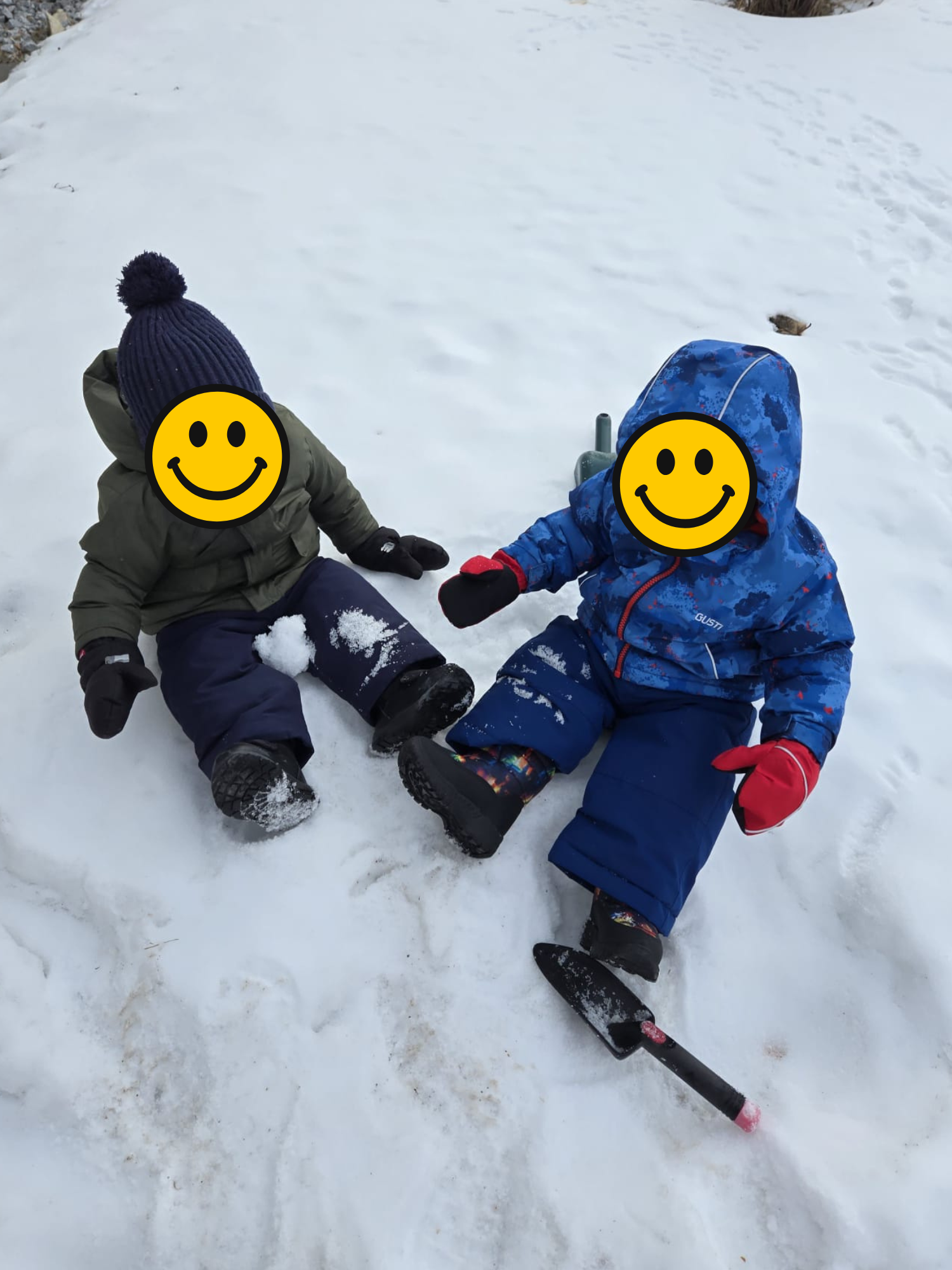 Two children sitting on snow with snowballs, wearing winter clothing and gloves, one with a navy blue hat and green jacket, the other with a blue camouflage jacket and red gloves, with a small snow shovel nearby.