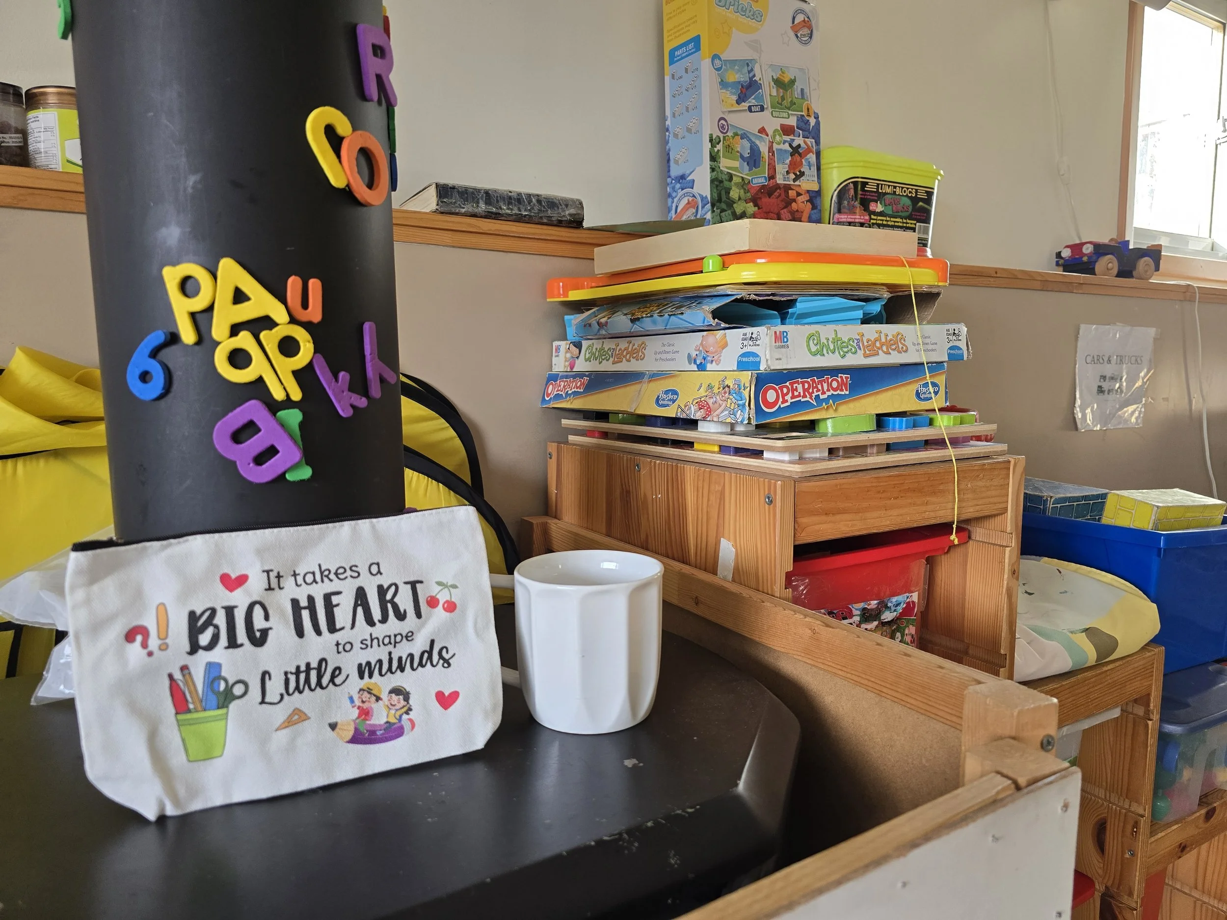 A classroom or playroom with various toys and educational materials. In the foreground, a black cylindrical object with colorful foam letters attached. In front of it, a white fabric bag with a heart and cartoon children illustration, and a white mug. Behind, a wooden tiered organizer with stacked board games including "Chutes and Ladders" and "Operation." There are additional toys and boxes on shelves and tables, with a window in the background.