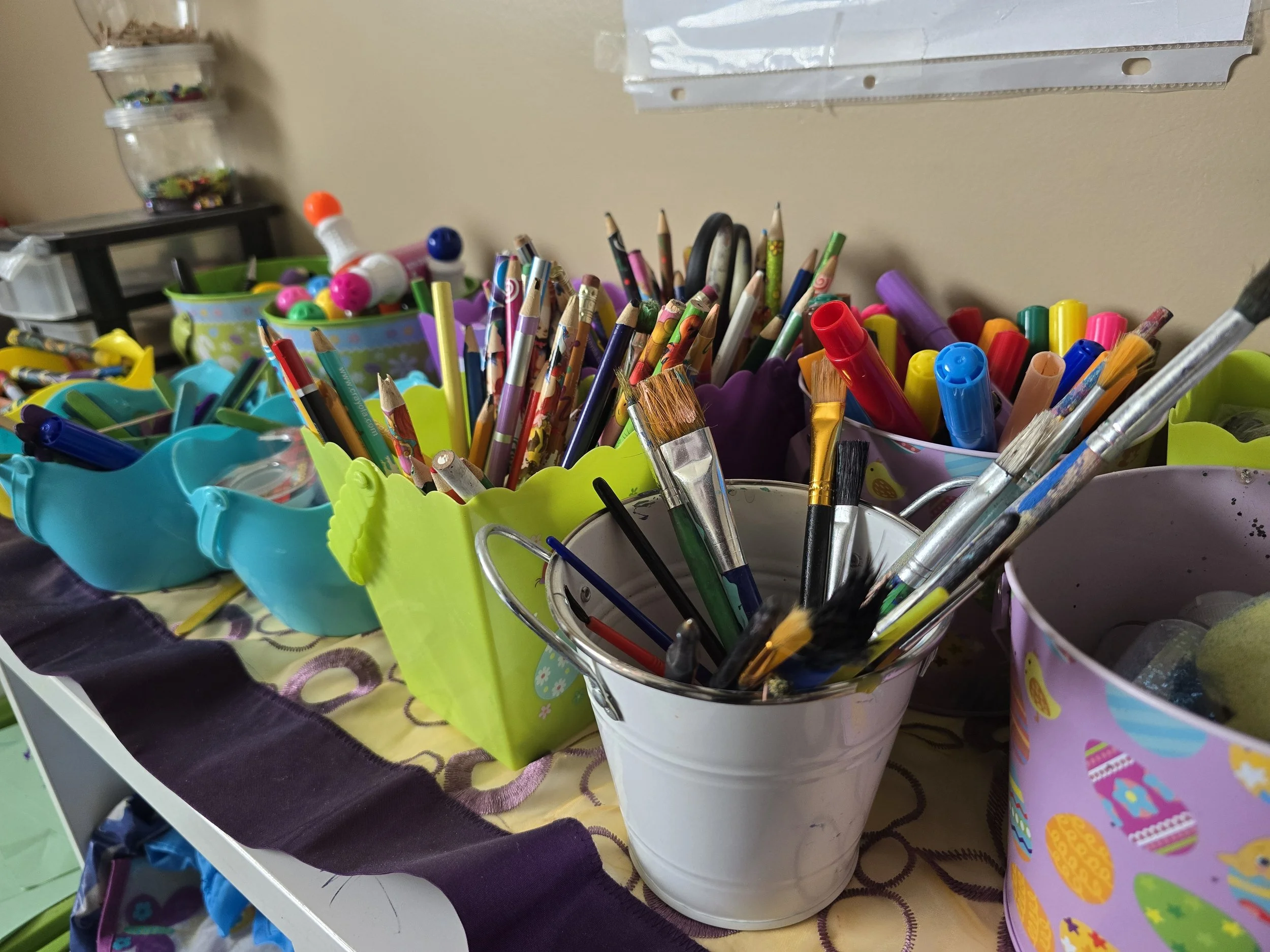 Collection of colorful pens, markers, and pencils in various containers on a table, with Easter-themed decorations in the background.