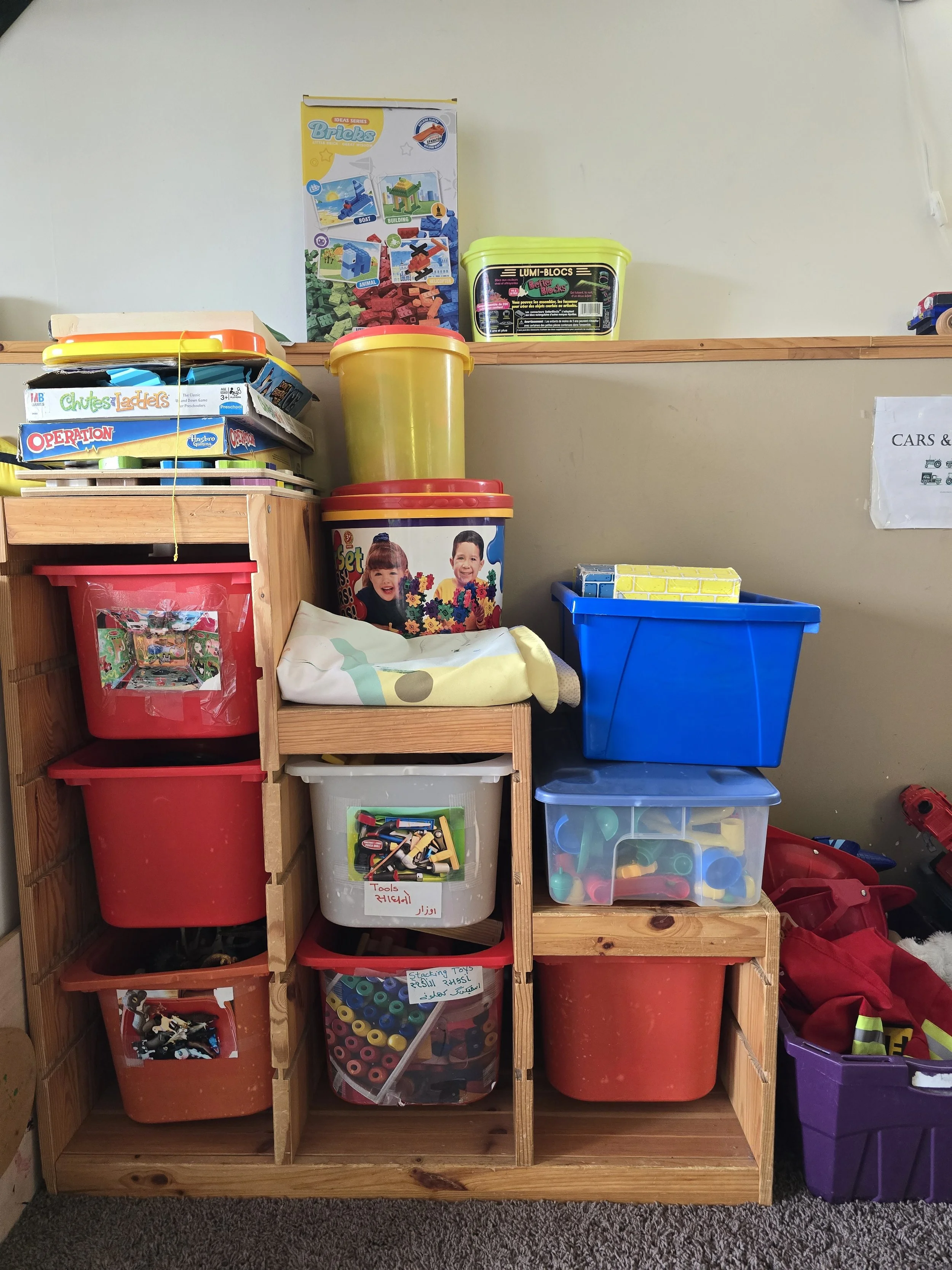 Storage area with various colorful plastic bins and containers filled with toys, including building blocks, toy tools, and vehicle figures. Some containers are labeled, and there is a box of Lego bricks on a wooden shelf.