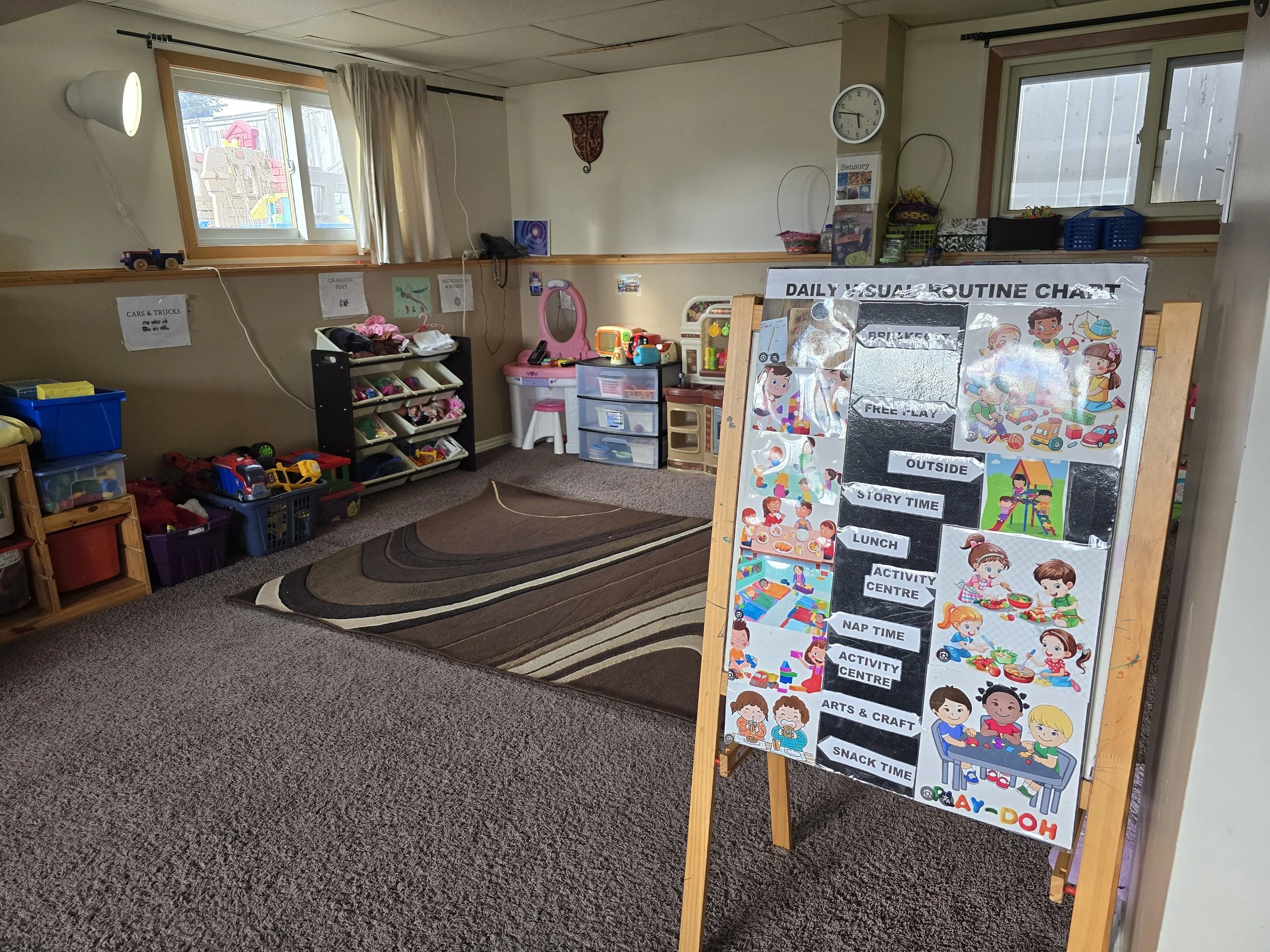 Indoor playroom with storage bins of toys, a play kitchen set, a rocking mirror, and a daily routine chart on an easel. The room has windows, curtains, and a carpeted floor.