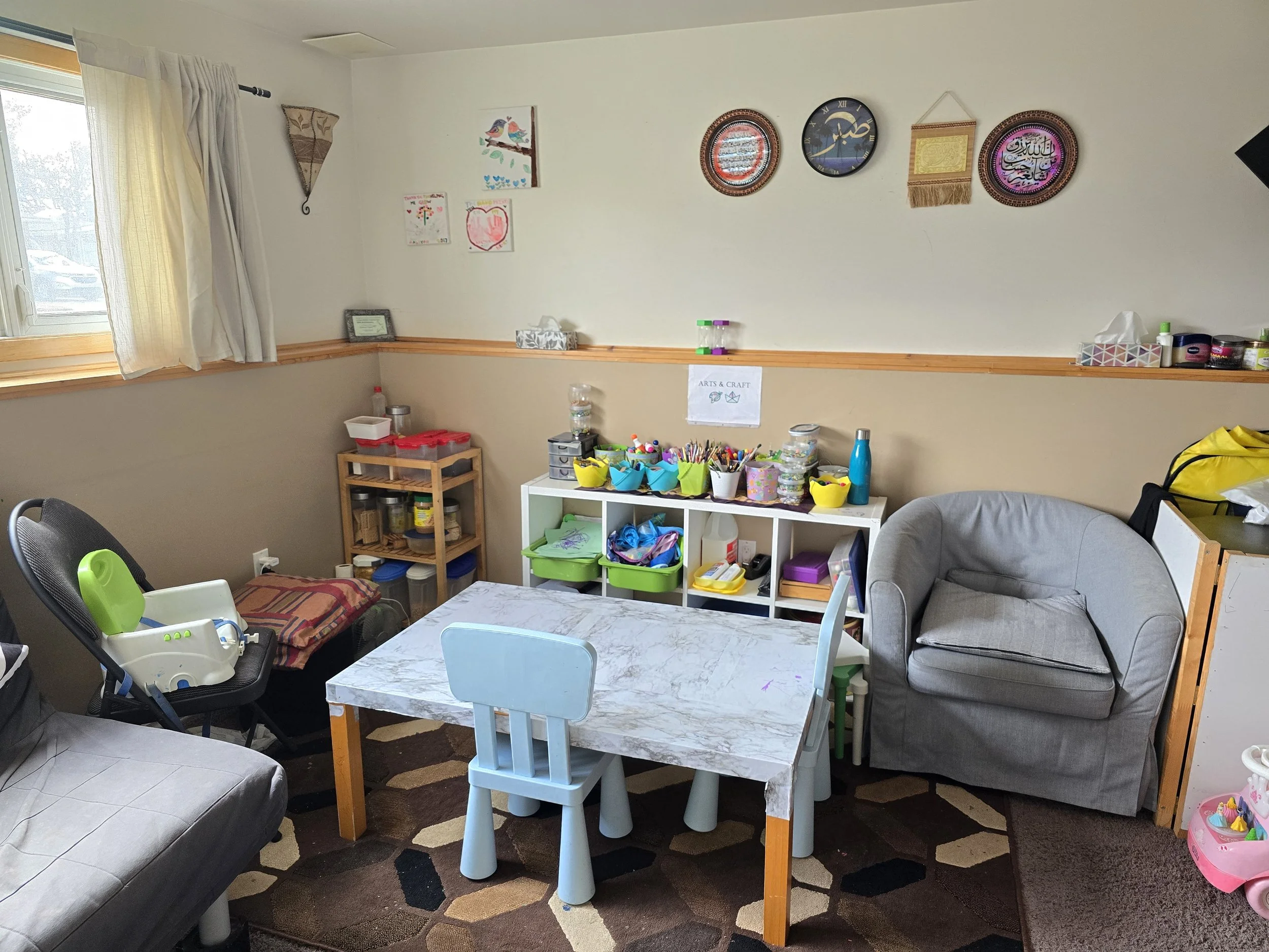 Child's playroom with a small table and chairs, a gray armchair, a window with curtains, and shelves containing toys, art supplies, and decorative wall hangings.