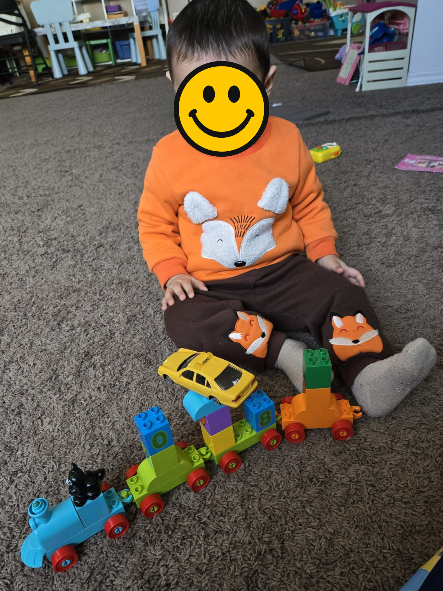 A young child sitting on a carpeted floor in a playroom with toy blocks and train set, wearing brown pants with fox designs and an orange sweatshirt with a fox face, with a smiley face sticker covering their face.