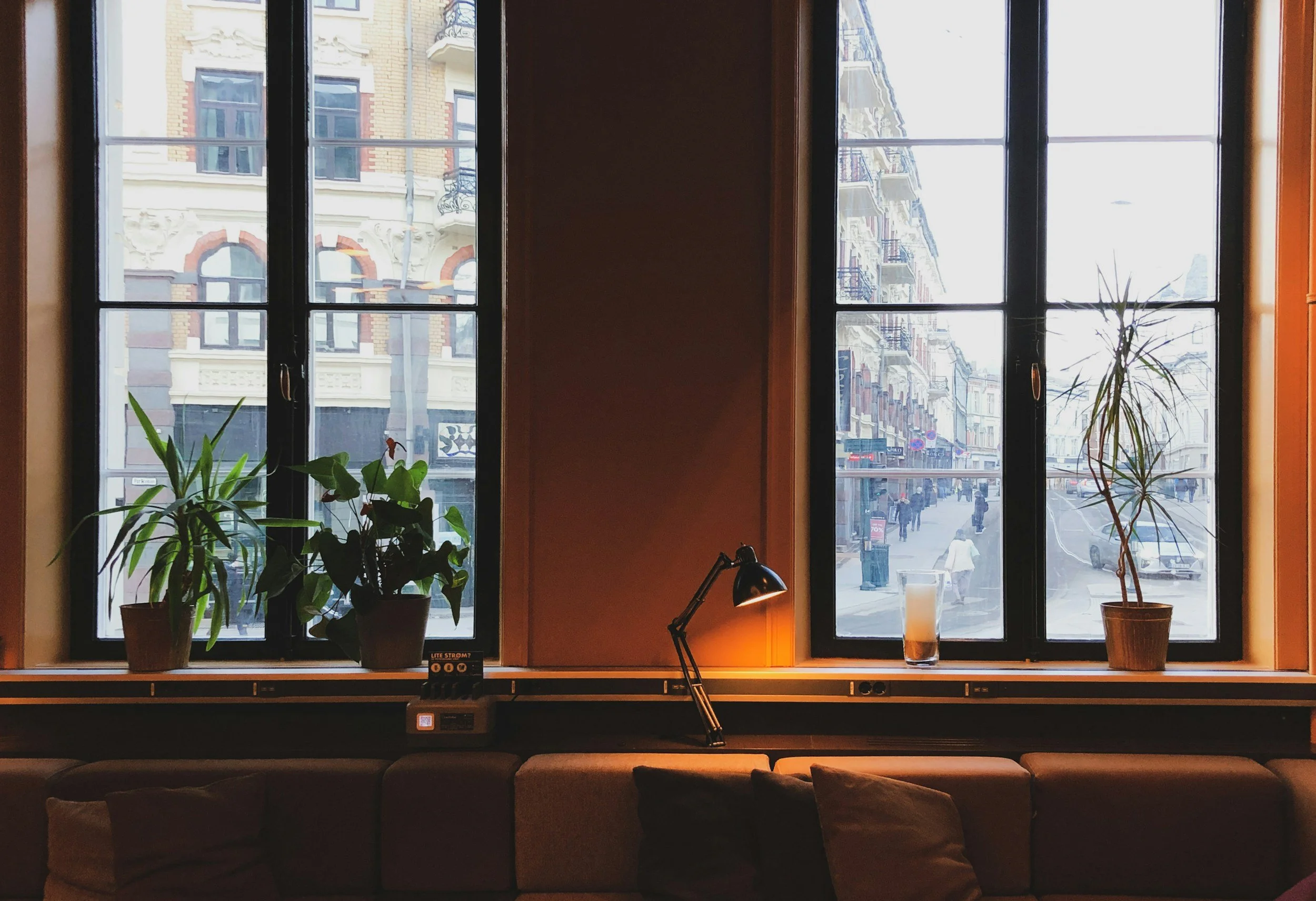 Bildet viser et rom med tre store vinduer som vender ut mot en gate i en by. Det er noen grønne planter på vinduskarmen, og et bordlampe står på en liten bordplate under vinduene.