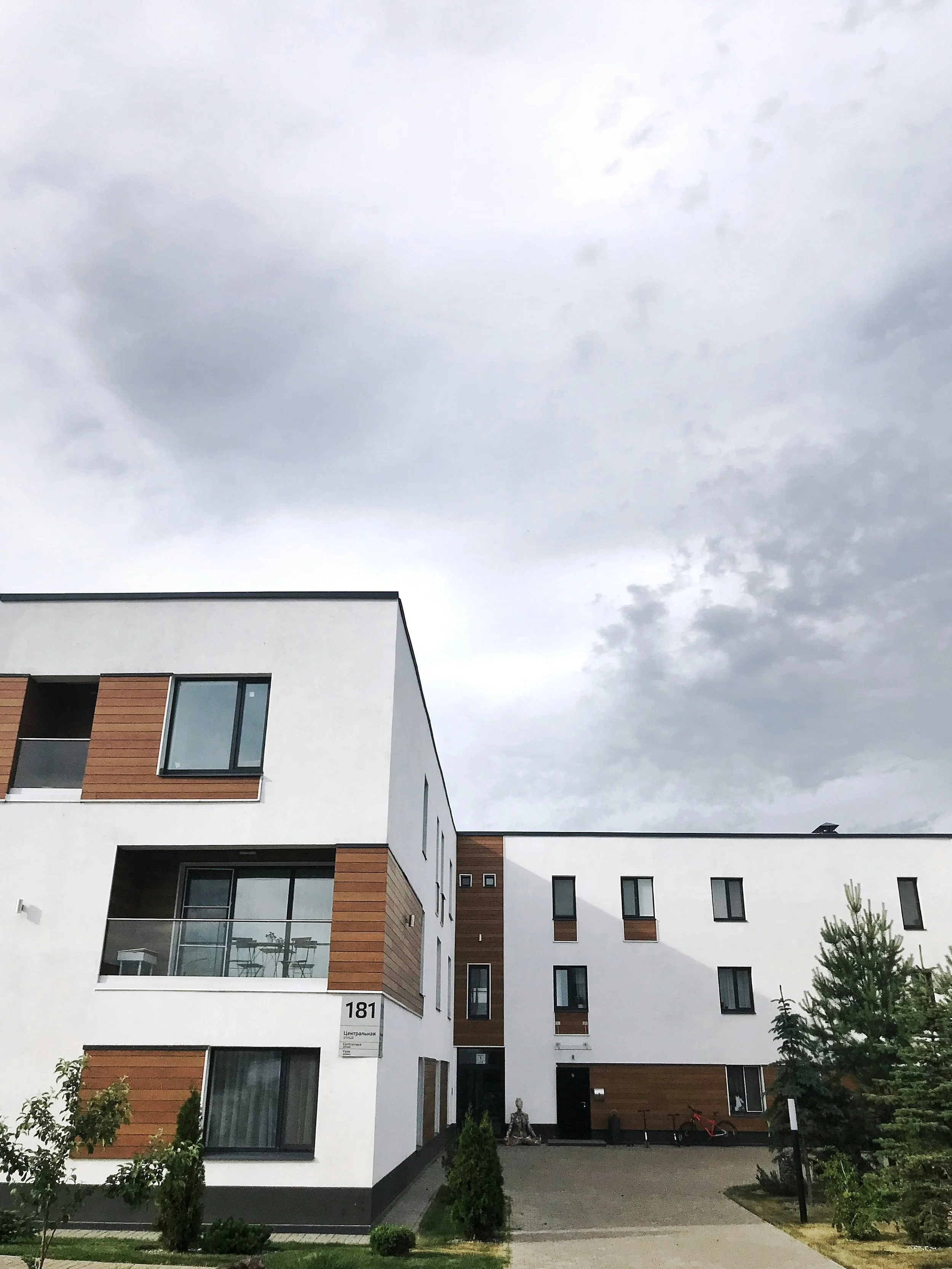 Fleksibelt bygningsdesign med hvit fasade, trepanel detaljer og store glassvinduer, omgitt av grøntområdet med busker og trær, under en overskyet himmel.