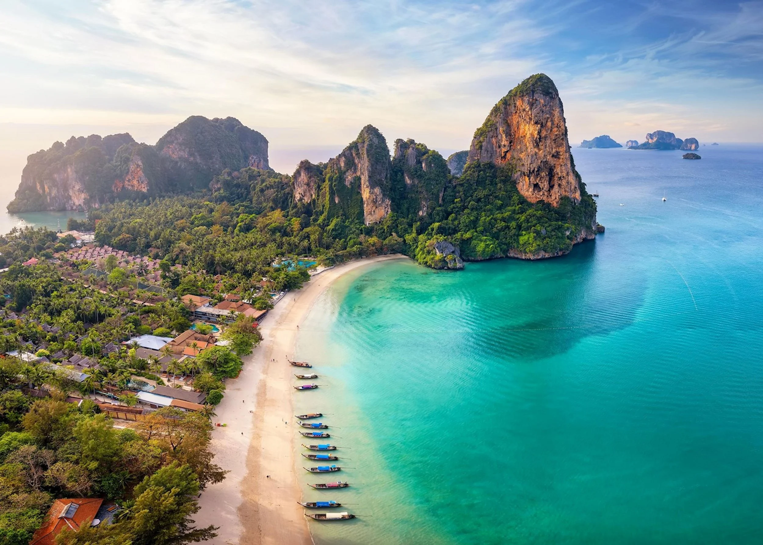 Aerial view of a tropical beach with turquoise water, surrounded by lush green hills and tall limestone cliffs. Several boats are anchored along the shoreline, with buildings and trees in the background under a partly cloudy sky.