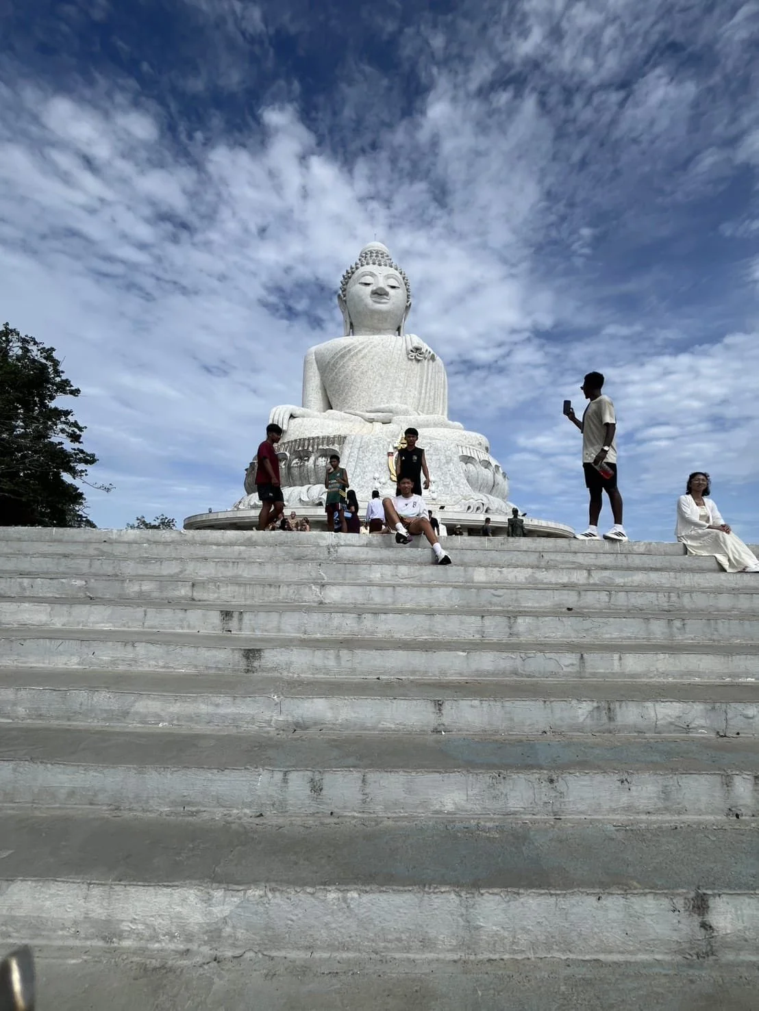 Large white Buddha statue on a stepped platform with several people sitting and standing around it, against a partly cloudy sky.