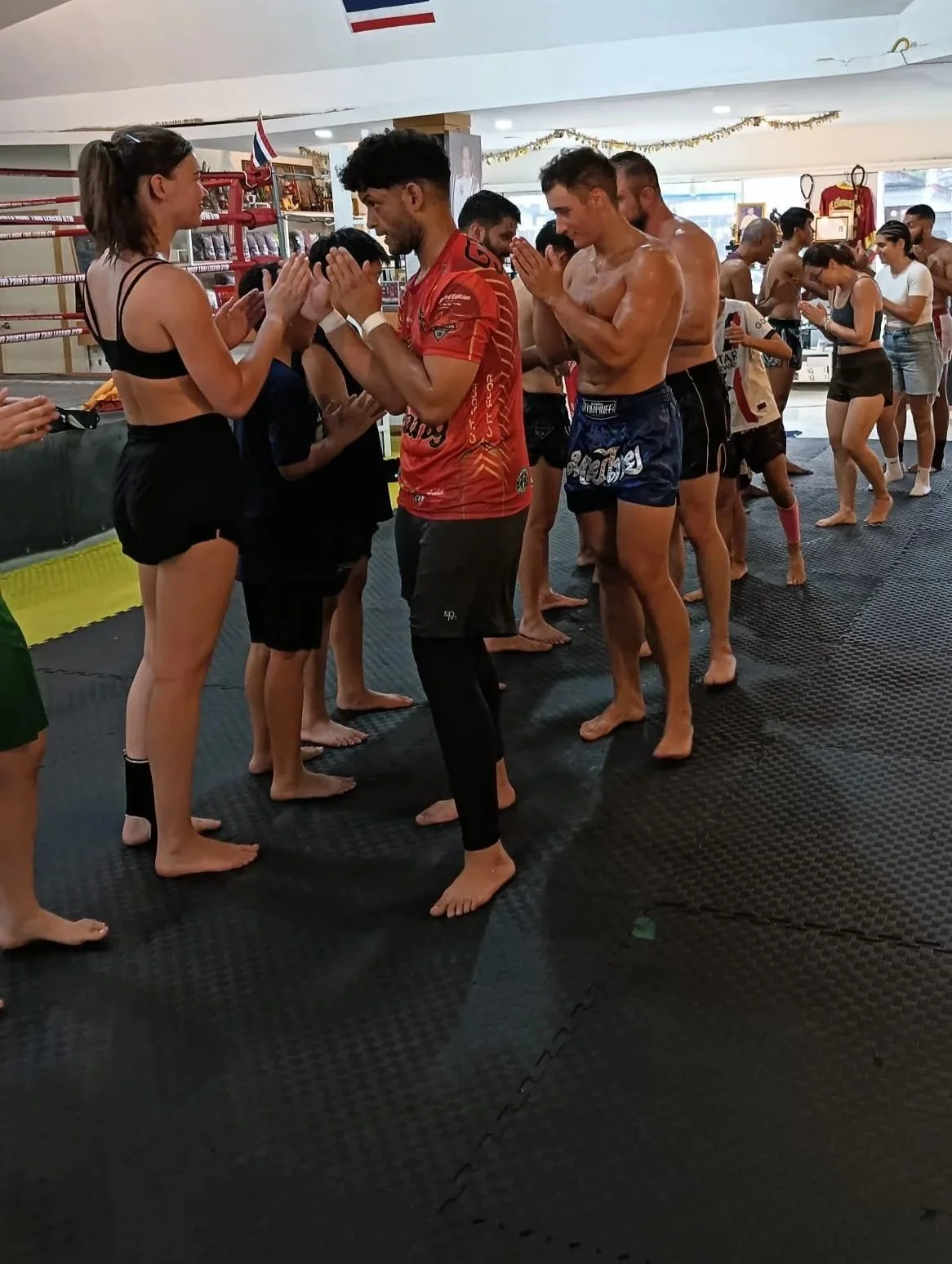 People practicing Thai martial arts or Muay Thai in an indoor gym, with some wearing traditional attire and others in athletic wear, engaging in a group training session.