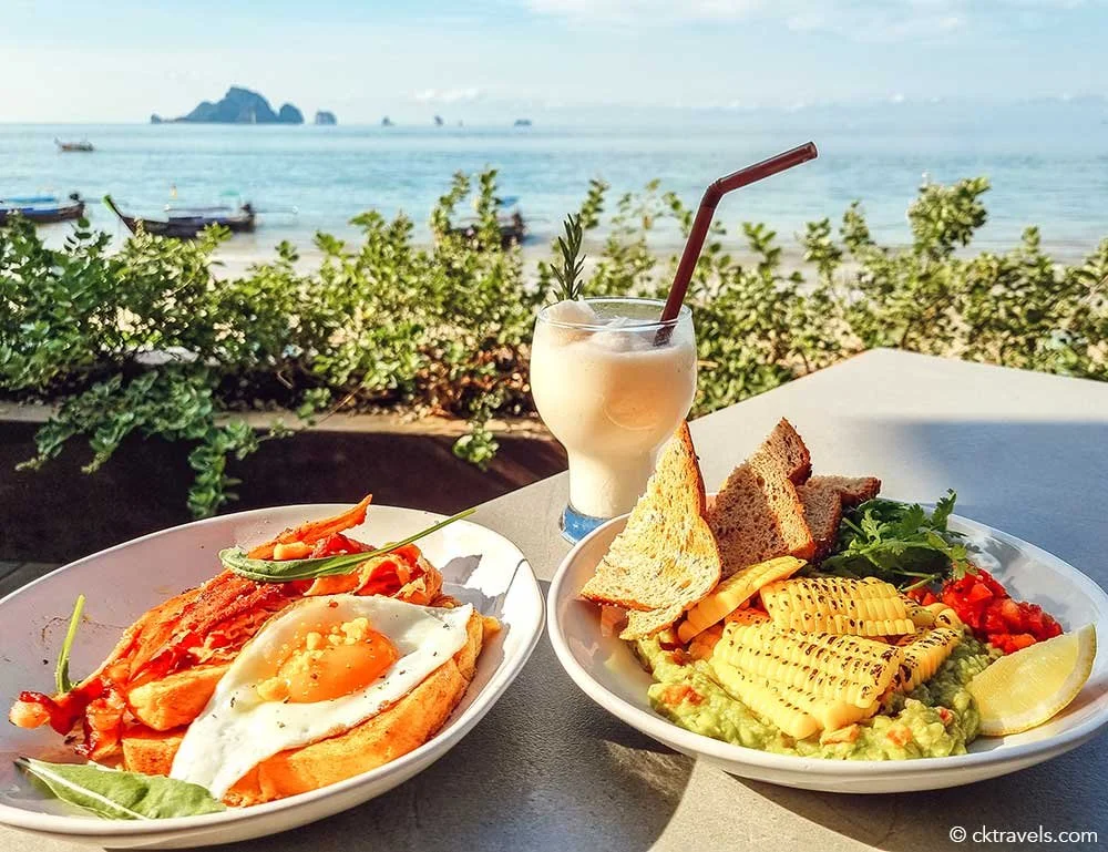 A seaside meal with two plates of food, a drink, and boats on the ocean in the background. One plate has eggs, vegetables, and bread; the other has grilled pineapple, avocado, toast, and salsa. There is a creamy beverage with a straw.