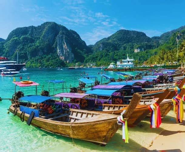 Boats docked on a tropical beach with turquoise waters, surrounded by lush green mountains under a clear blue sky.